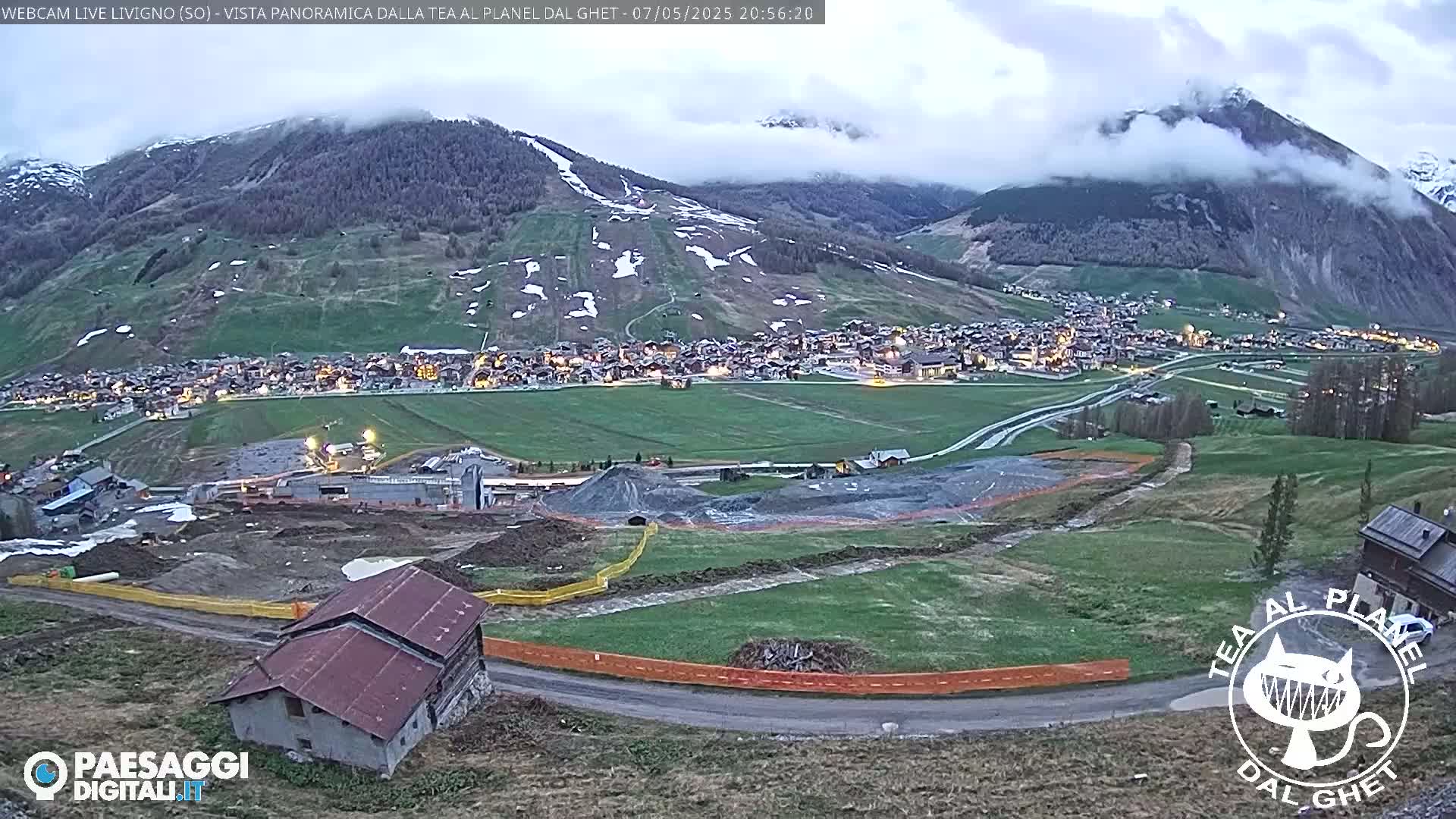 A mountain village at dusk, partially shrouded in fog, with a construction site in the foreground and snow-capped mountains in the background.