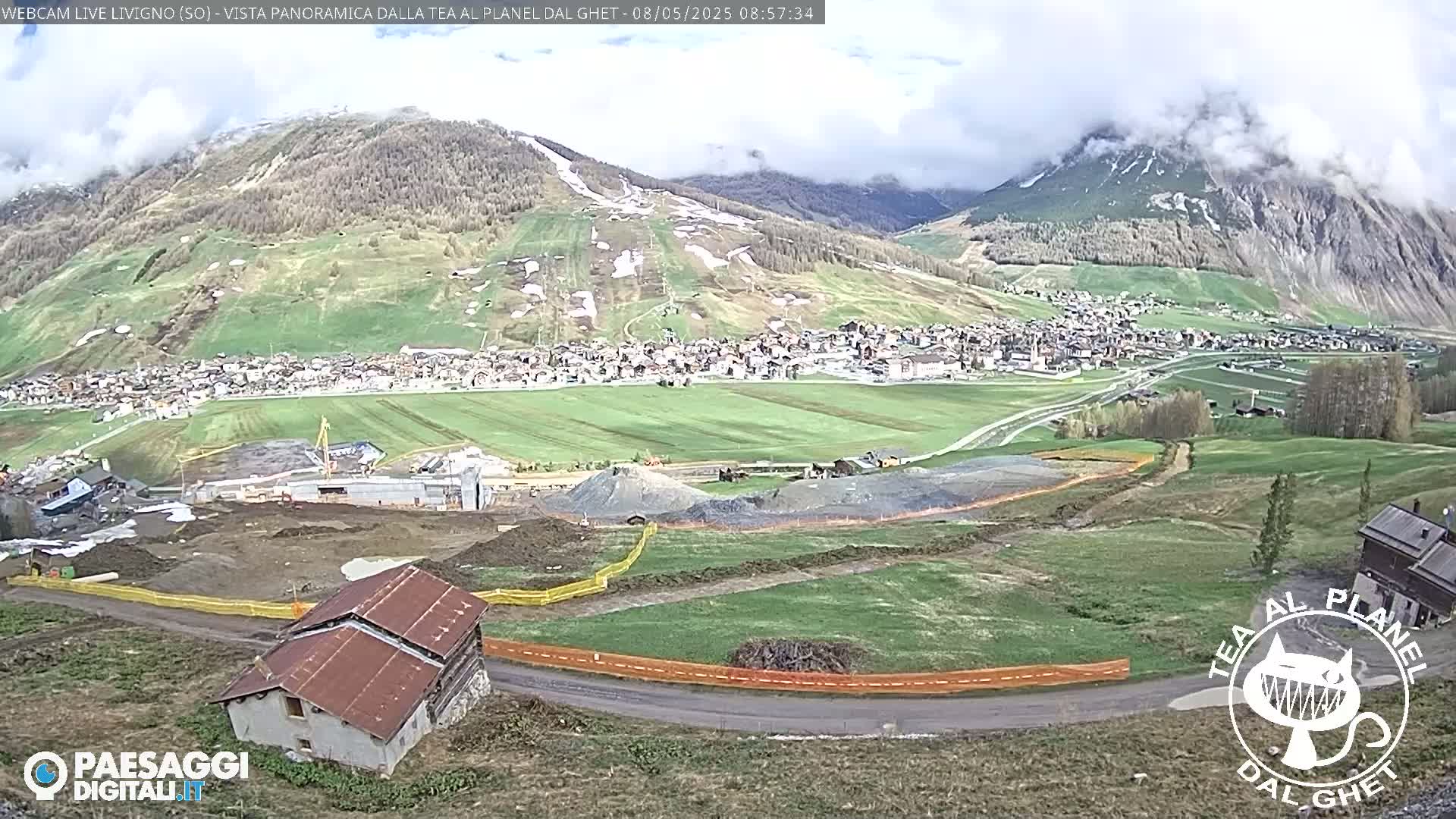 A partly cloudy day reveals a mountain village nestled in a valley, with construction underway near a small house and  patches of snow visible on the surrounding hillsides.