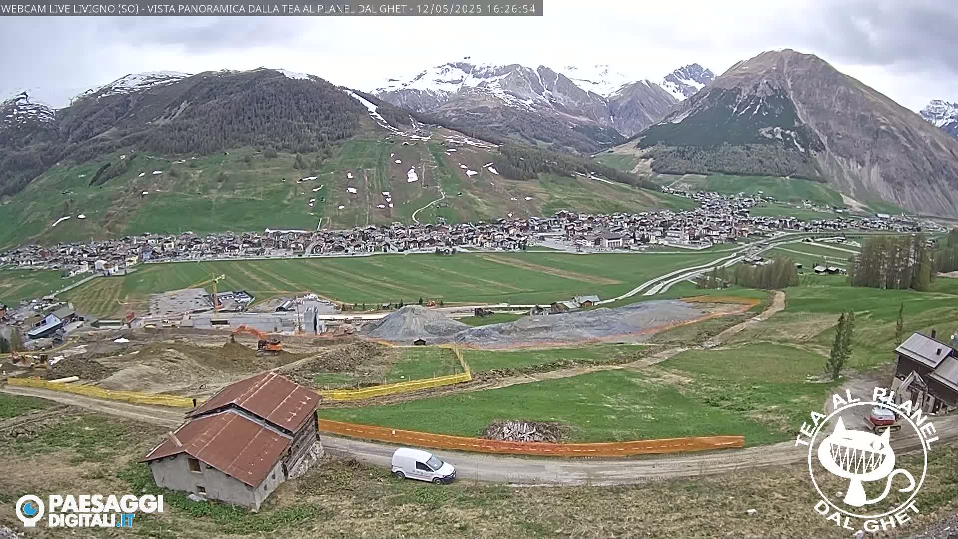 A partly cloudy day reveals a mountain village nestled in a valley, with construction activity visible in the foreground and snow-capped mountains in the background.