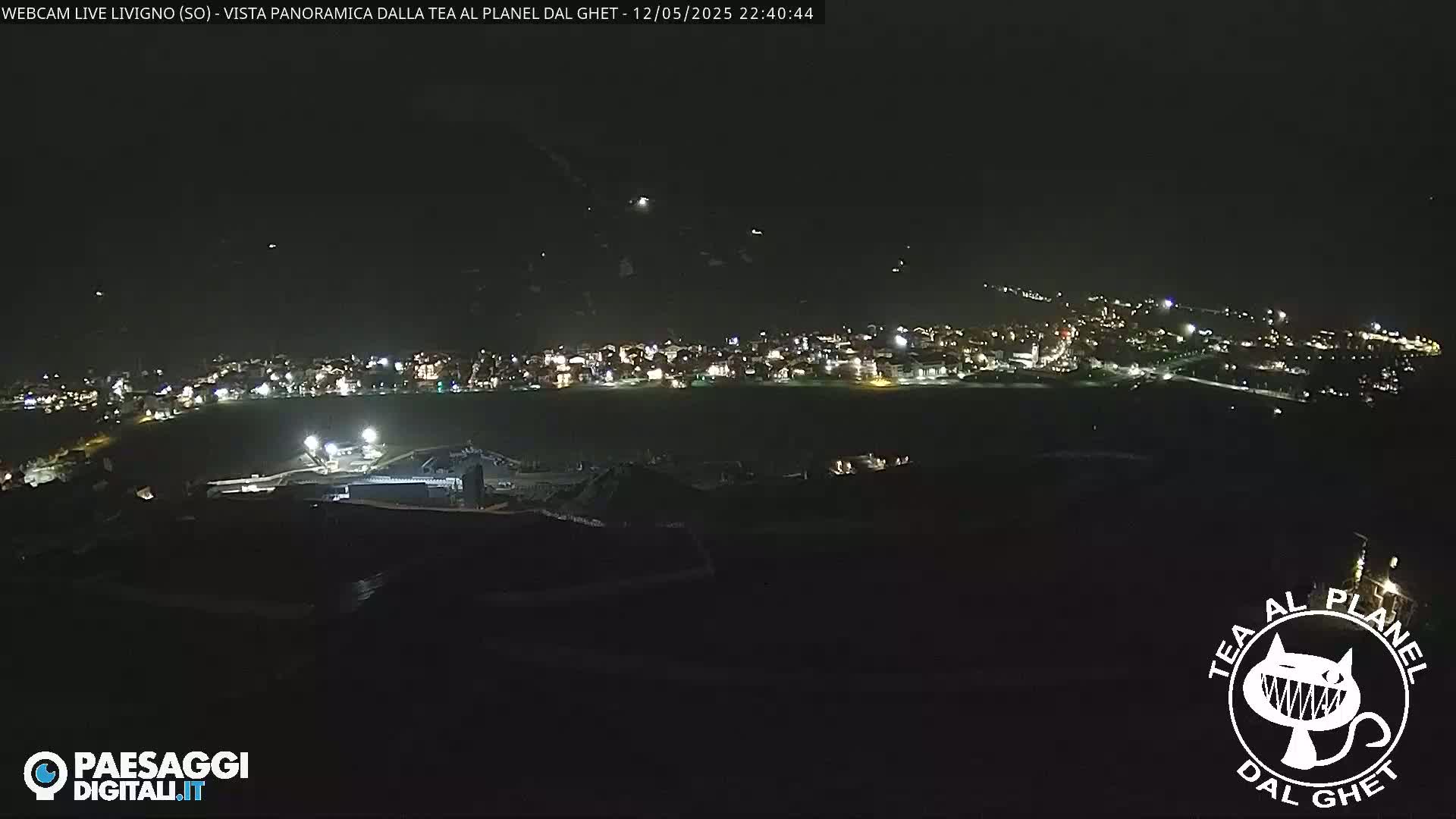 A nighttime panoramic view of a town illuminated by lights, situated below a dark mountainside under a clear, dark sky.