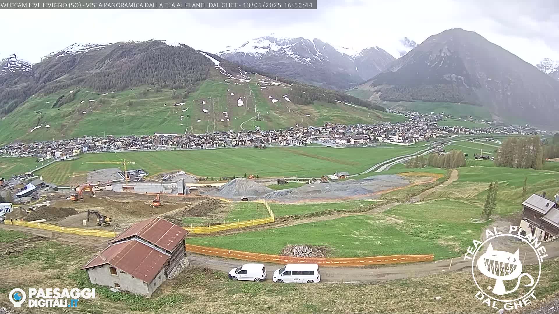 A panoramic view of a town nestled in a valley surrounded by green hills and snow-capped mountains under an overcast sky shows construction activity near a road and several vehicles.