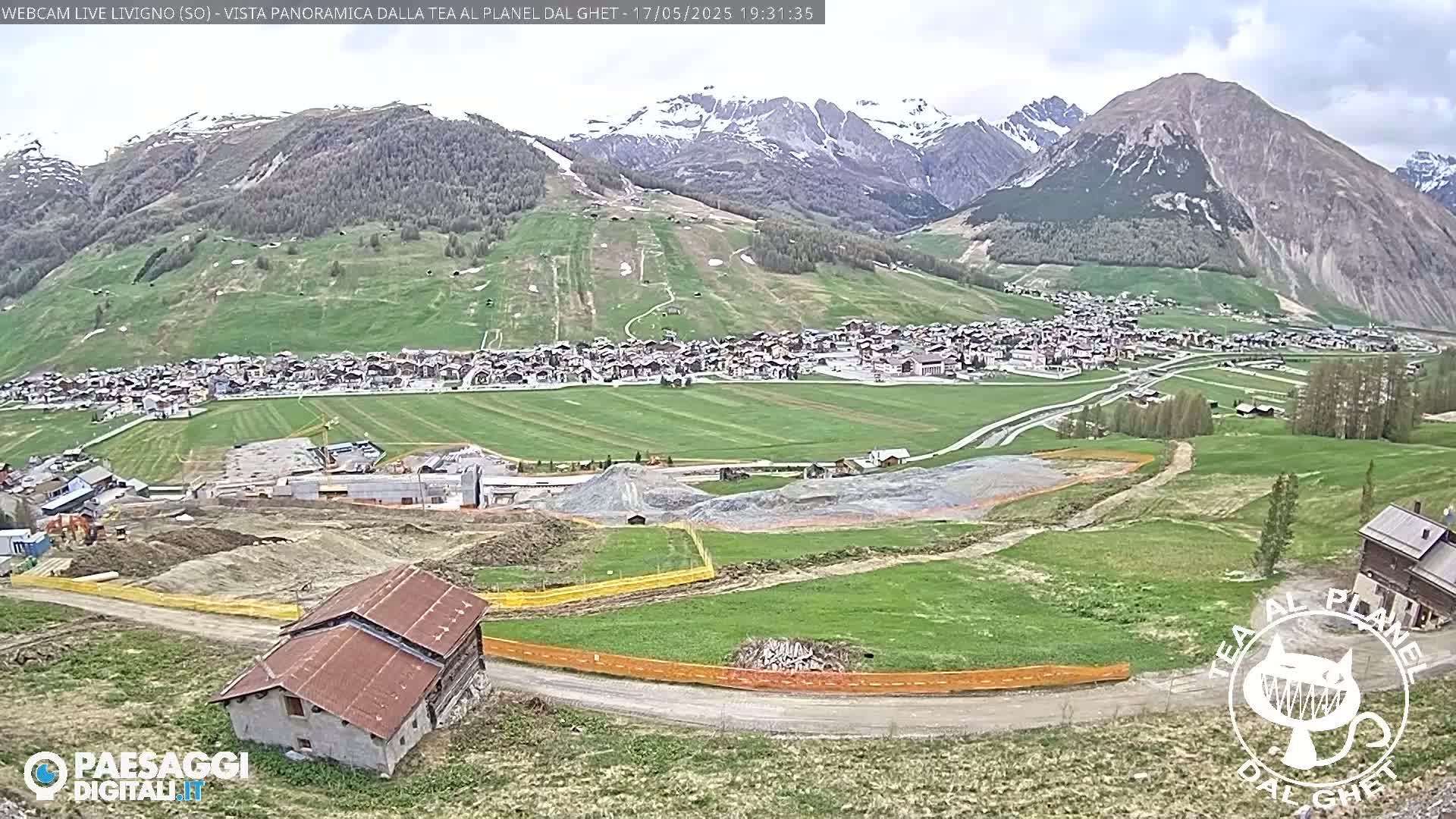 A mountain village nestled in a valley below snow-capped mountains under a partly cloudy sky, with ongoing construction visible in the foreground.