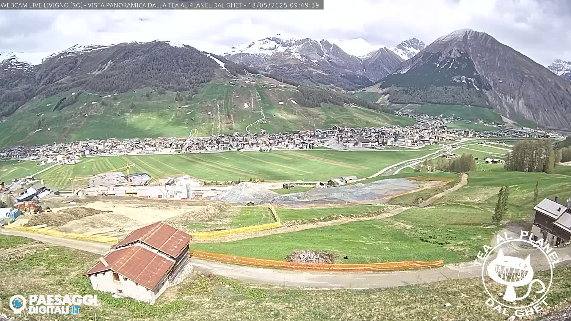 A mountain town nestled in a valley, with snow-capped mountains in the background under partly cloudy skies, shows ongoing construction near a small house.