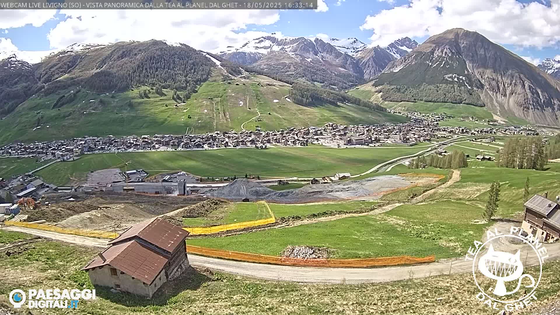 A mountain village nestled in a valley below snow-capped mountains on a partly cloudy day, with construction or land clearing visible in the foreground.