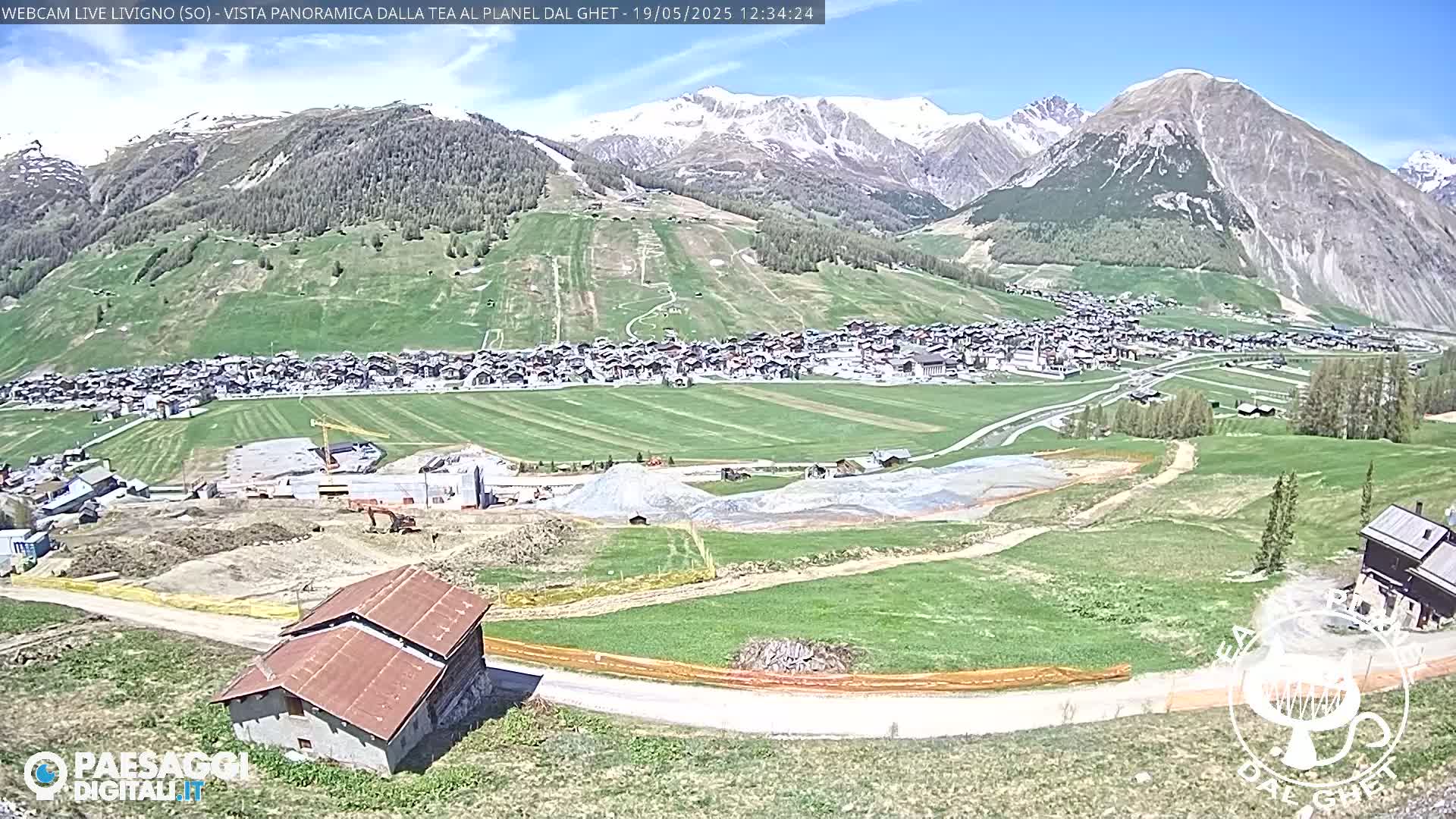 A sunny mountain village nestled in a valley with green fields and a construction site, snow-capped mountains in the background.