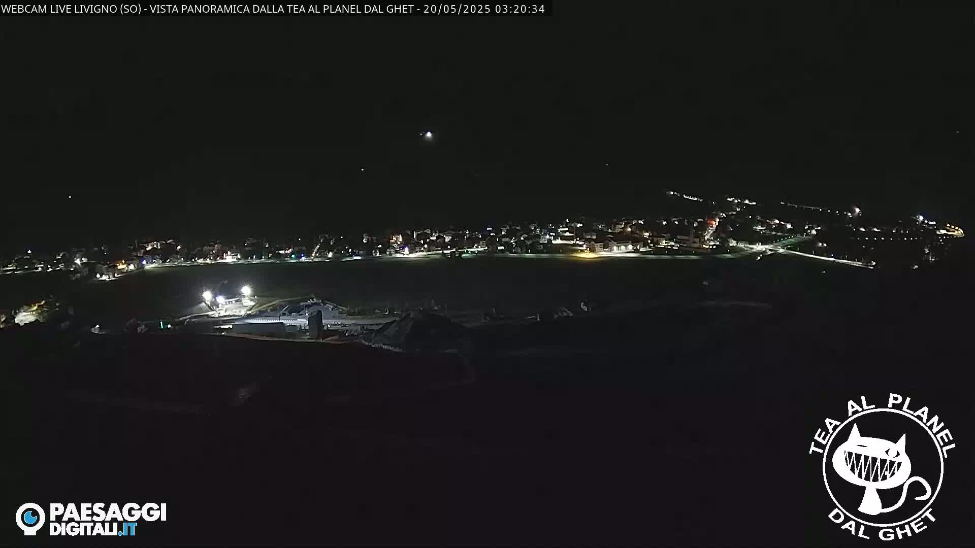 A nighttime, high-angle view shows a small town illuminated by lights, situated in a dark, possibly mountainous area.