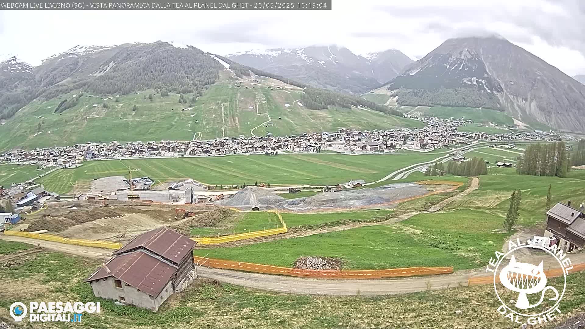 A panoramic view of a town nestled in a valley surrounded by green hills and mountains under an overcast sky, with construction visible in the foreground.