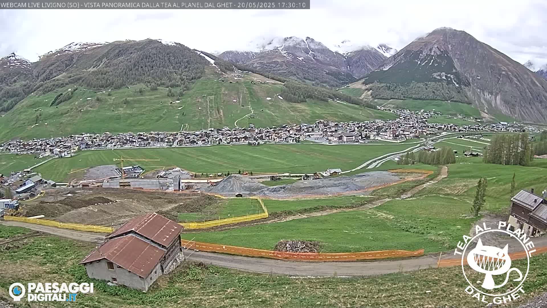 A partially cloudy day reveals a village nestled in a green valley surrounded by mountains, with some ongoing construction visible in the foreground.