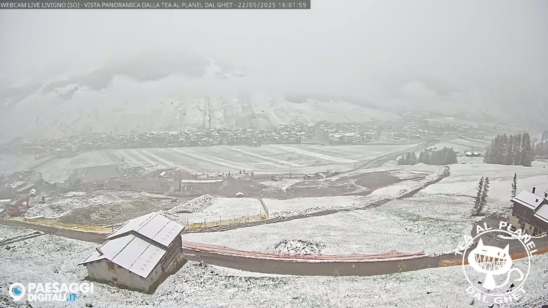 A snow-covered landscape featuring a village in the distance, construction sites, and a few buildings closer to the viewer, under a light snowfall.