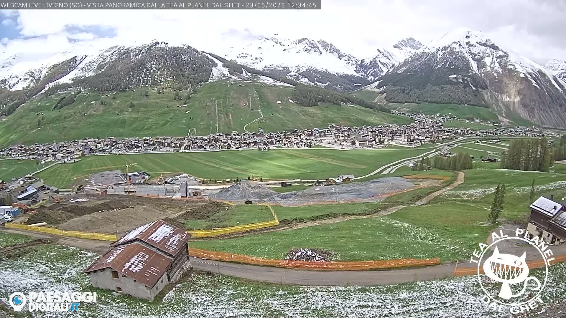 A snow-dusted mountain range overlooks a village nestled in a valley of green fields and construction sites under a partly cloudy sky.
