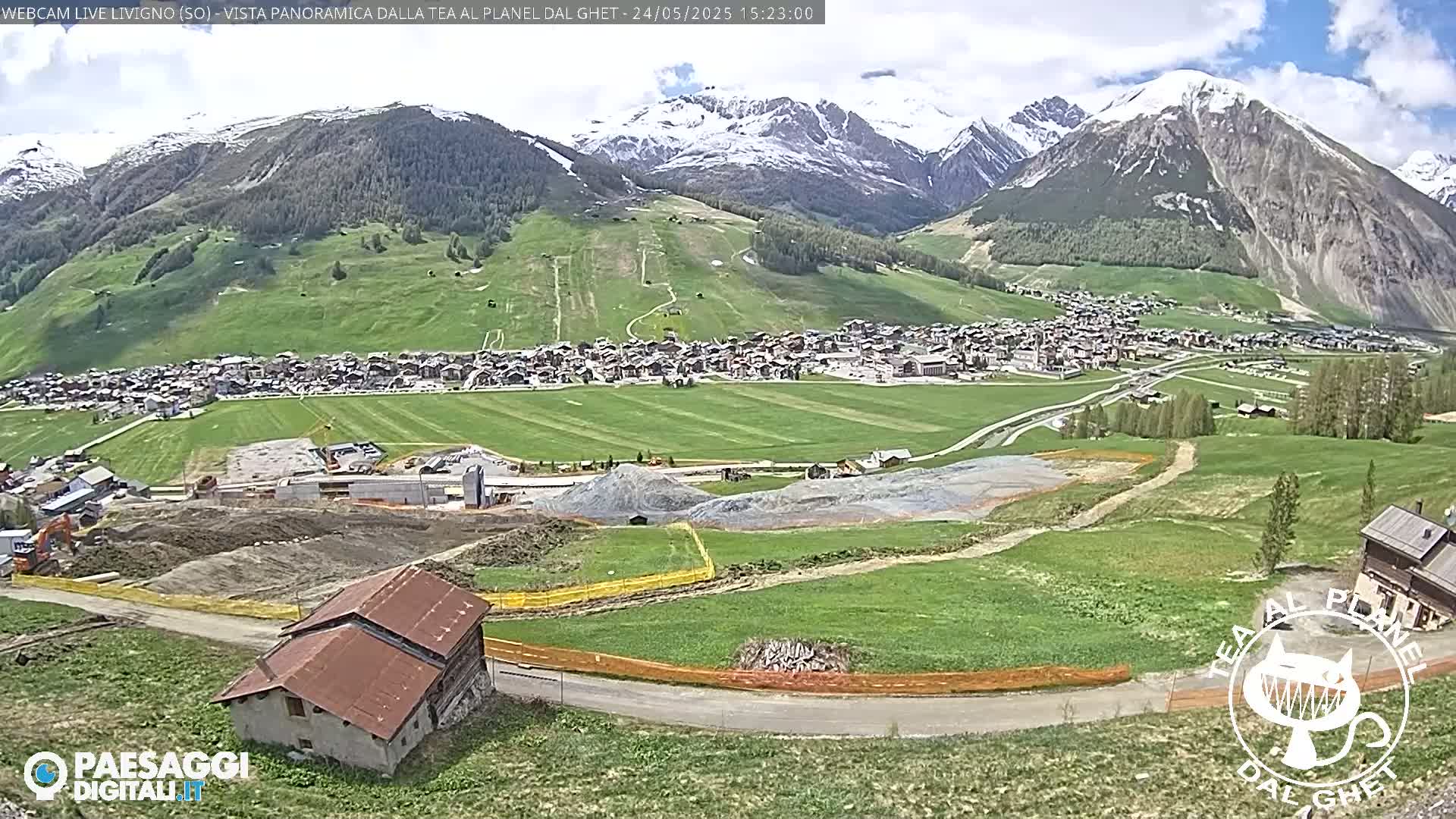 A partly cloudy day reveals a mountain village nestled in a valley, with snow-capped mountains in the background and construction work visible in the foreground.
