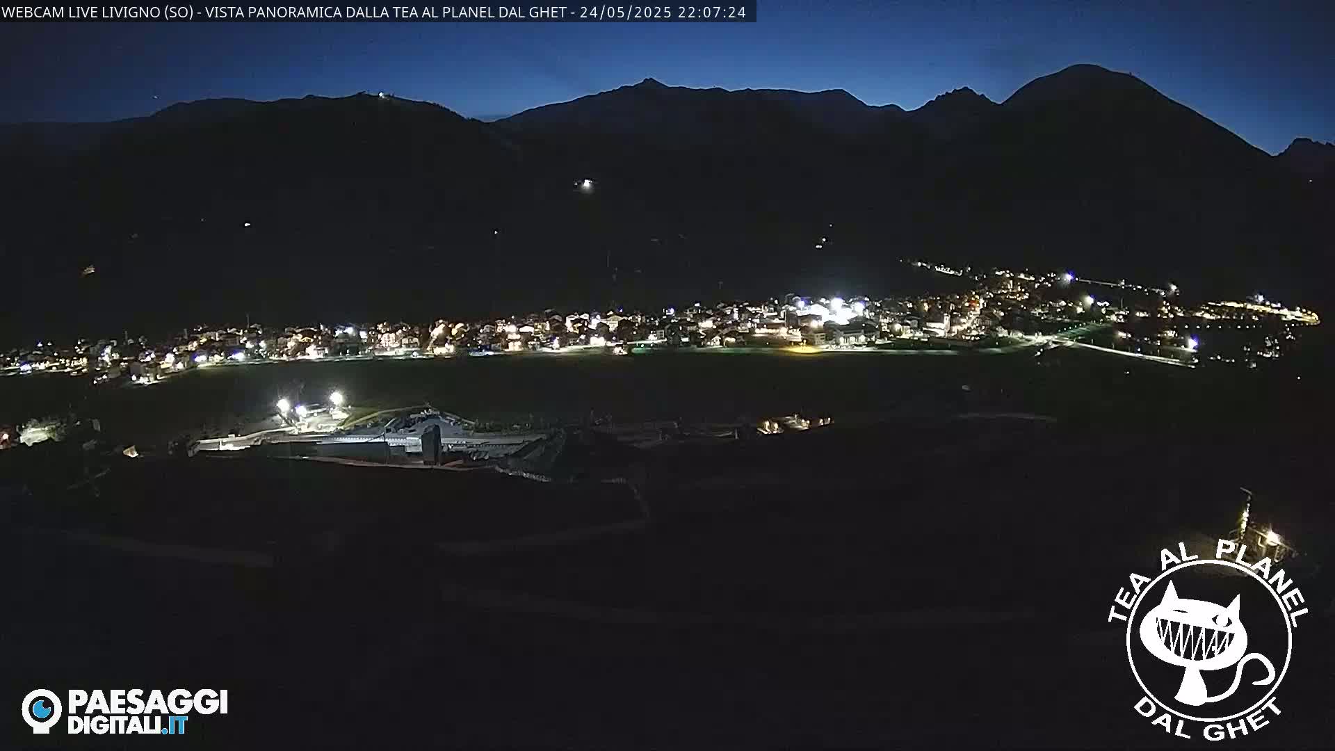 A nighttime, panoramic view of a small town nestled in a valley between dark mountains under a clear, dark sky.