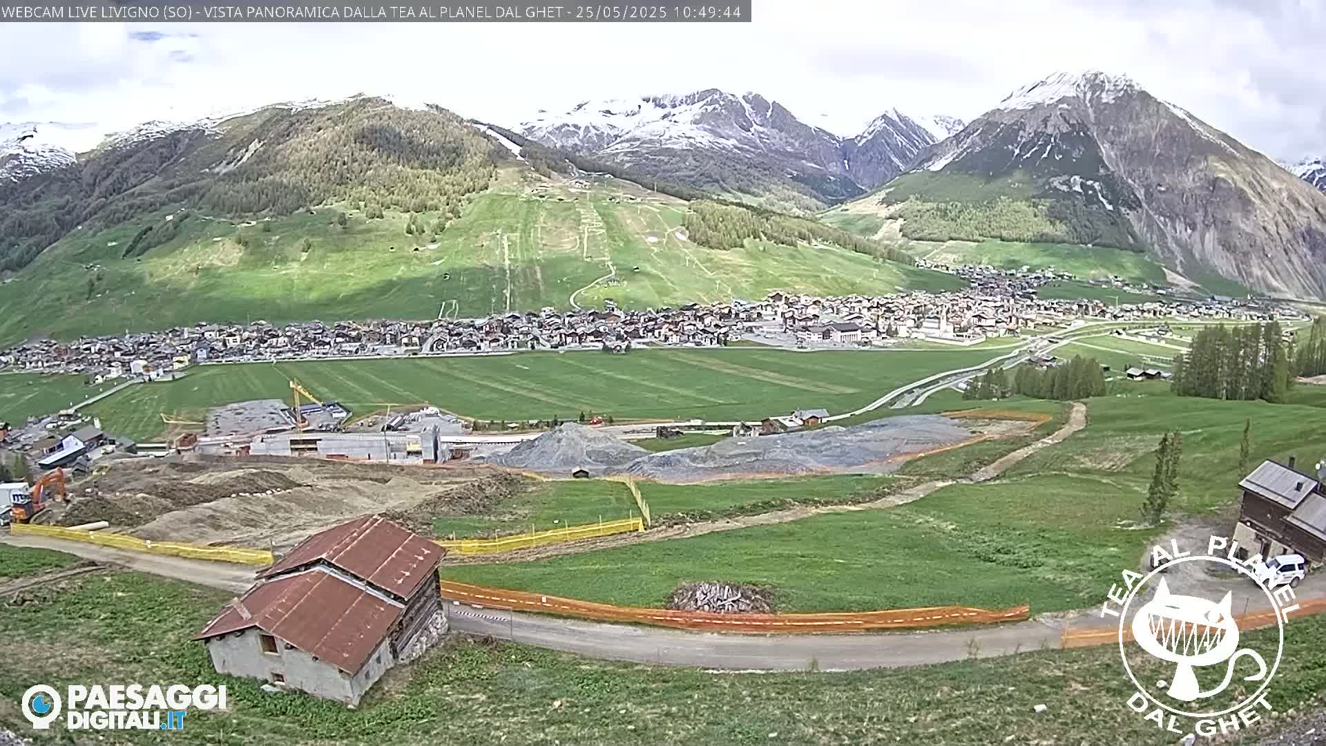 A partially cloudy day reveals a mountain village nestled in a green valley, with construction visible in the foreground and snow-capped mountains in the background.