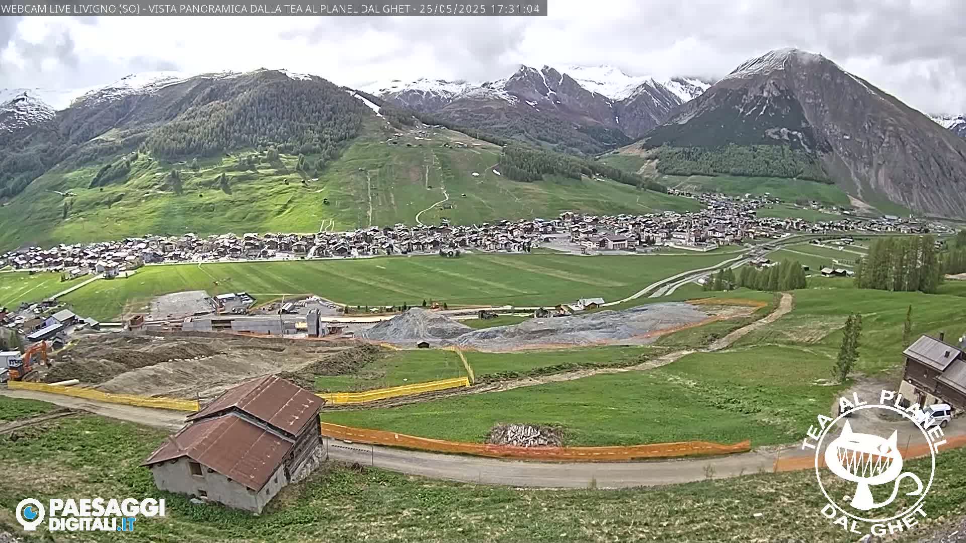 A mountain village nestled in a valley under a cloudy sky, with construction activity visible in the foreground and snow-capped mountains in the background.