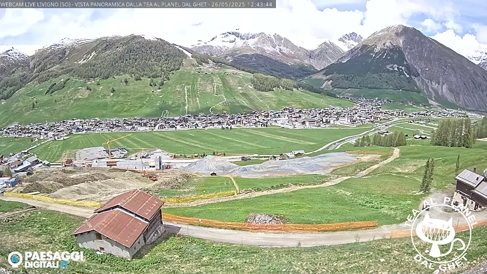 A partially cloudy day reveals a mountain town nestled in a valley, with green hills and construction visible in the foreground.