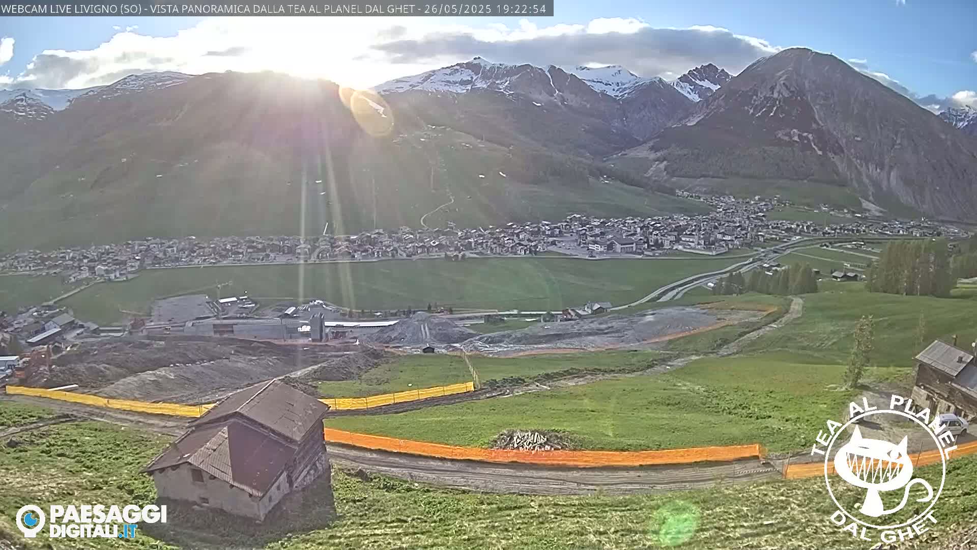 A mountain village nestled in a valley beneath snow-capped mountains, bathed in the late afternoon sun, with partially cloudy skies.