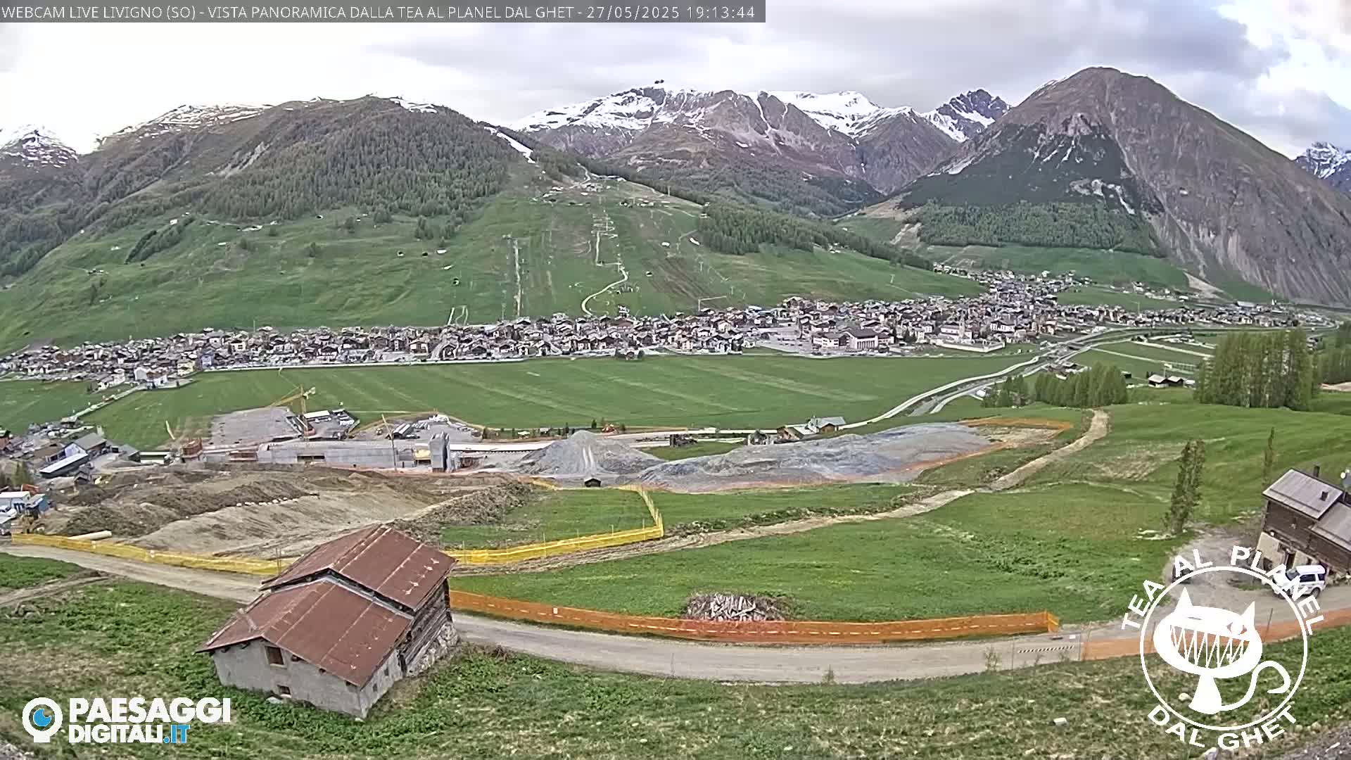 A mountain village nestled in a valley, with snow-capped mountains in the background and partly cloudy skies, shows construction activity in the foreground.