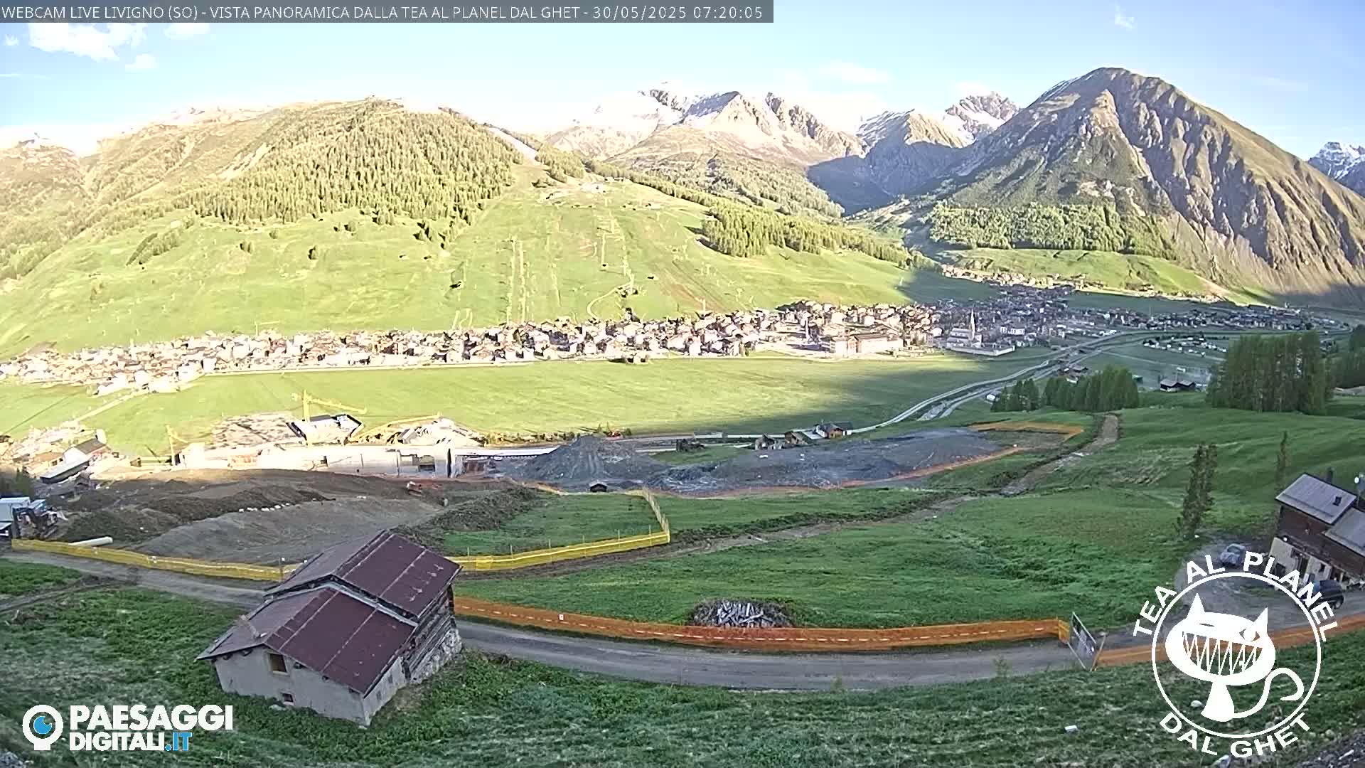 A sunny mountain valley town is viewed from above, showing a construction site in the foreground and a road curving down to a small building.