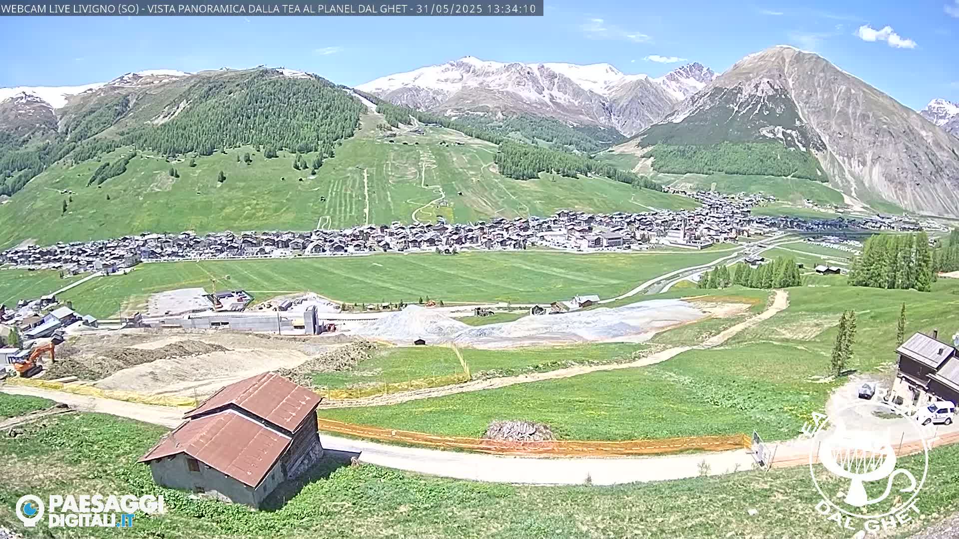 A sunny view of a town nestled in a green valley, surrounded by mountains with some snow, and a construction site in the foreground.