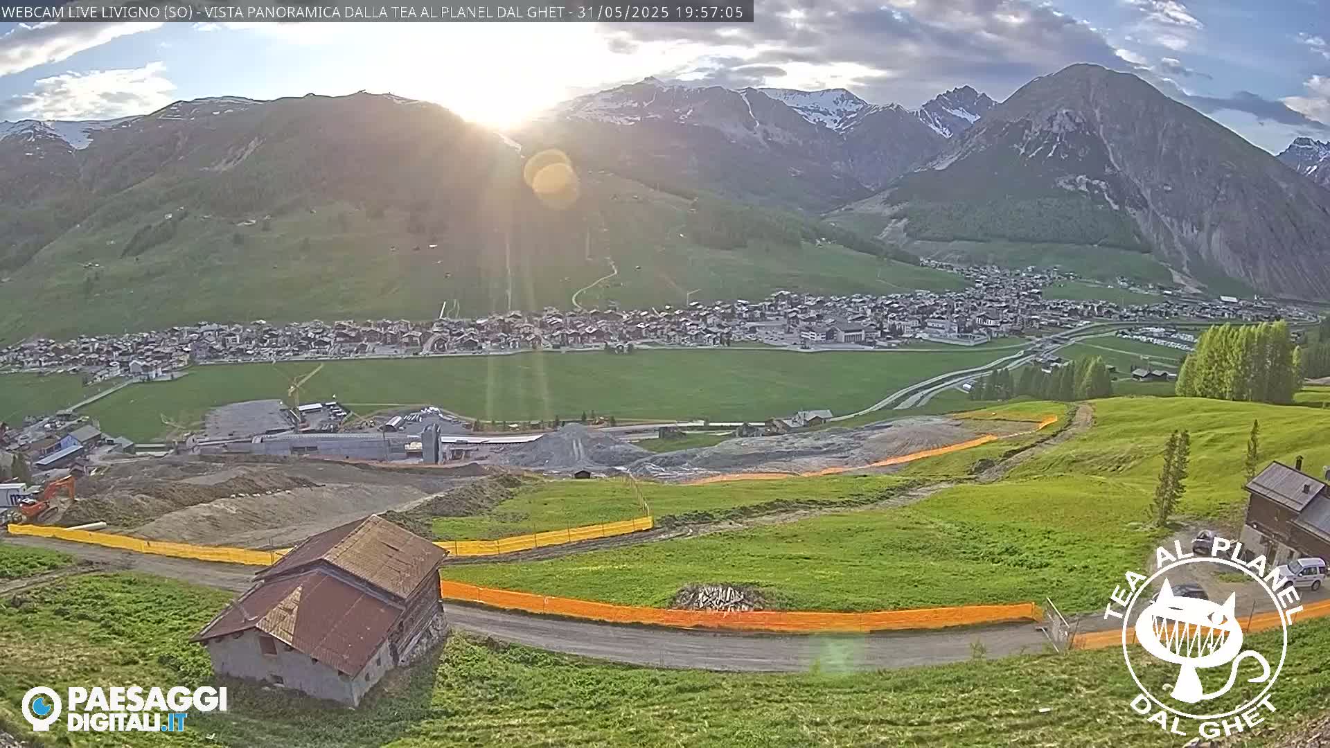 A sunlit mountain town nestled in a valley below snow-capped peaks, with a construction site and a small building in the foreground, under mostly clear skies.