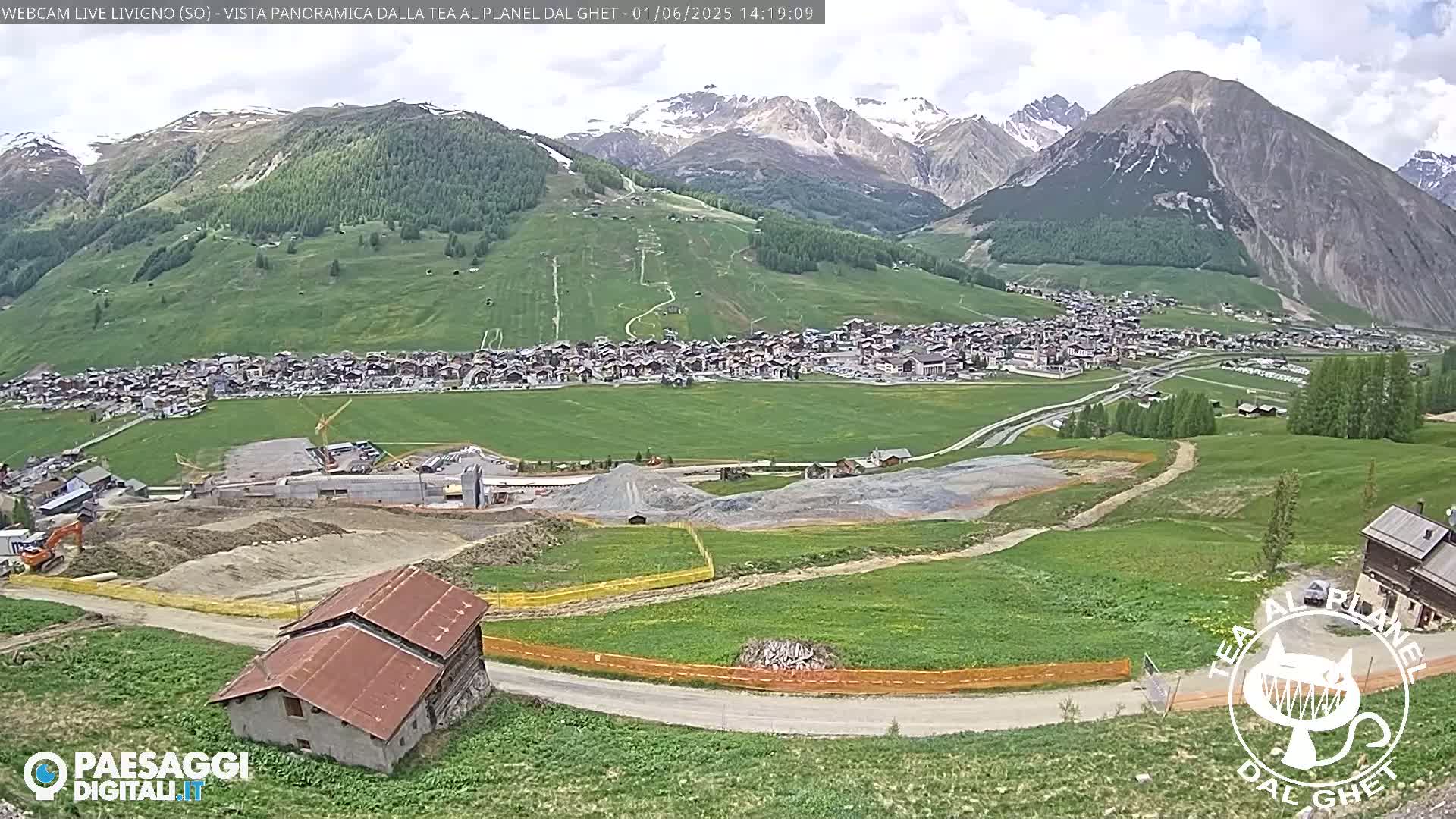 A partly cloudy day reveals a mountain village nestled in a green valley, with construction activity visible in the foreground and snow-capped mountains in the background.