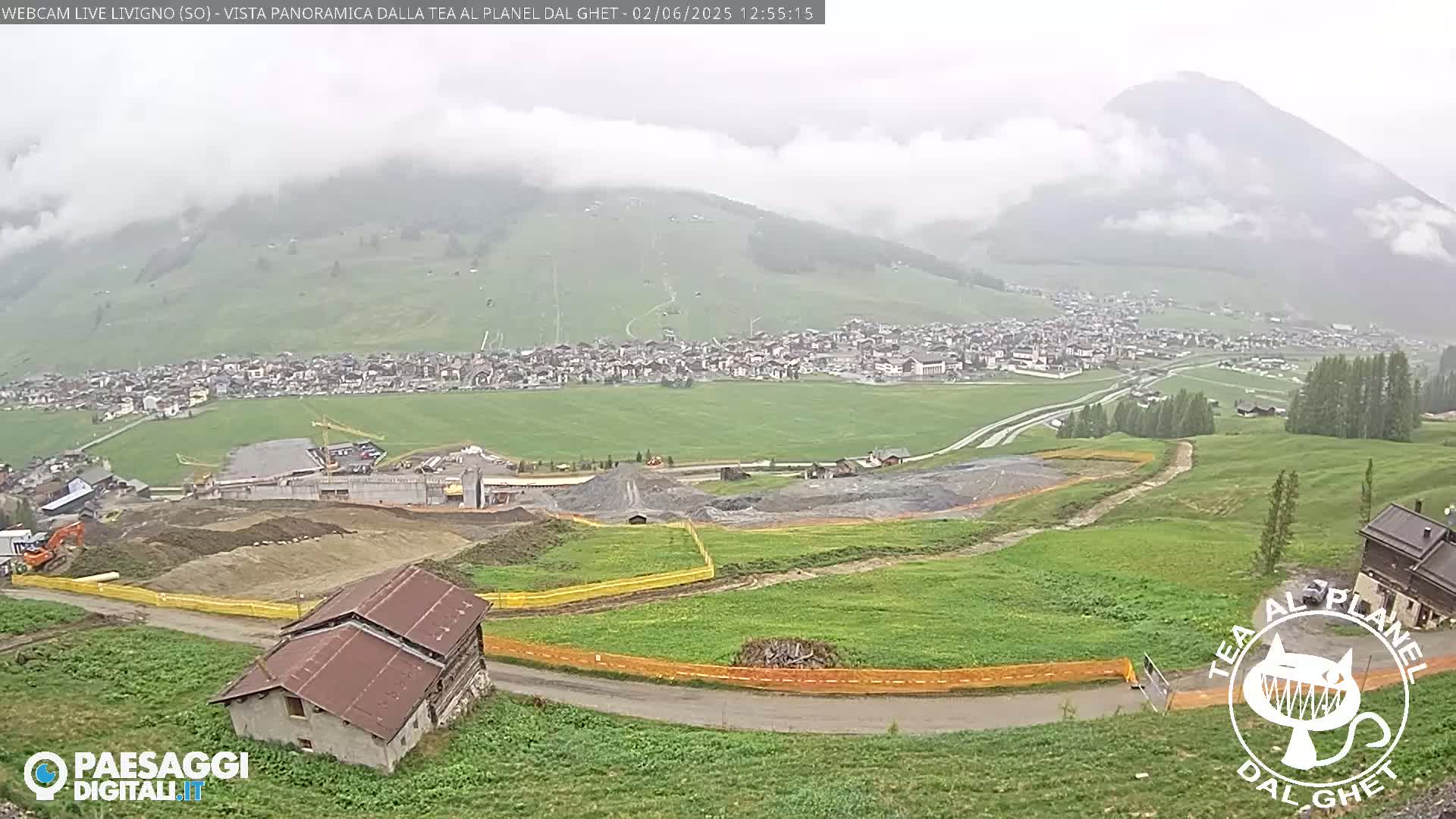 A mountain village is visible in the distance beneath a cloudy sky, with construction and excavation work underway in the foreground.