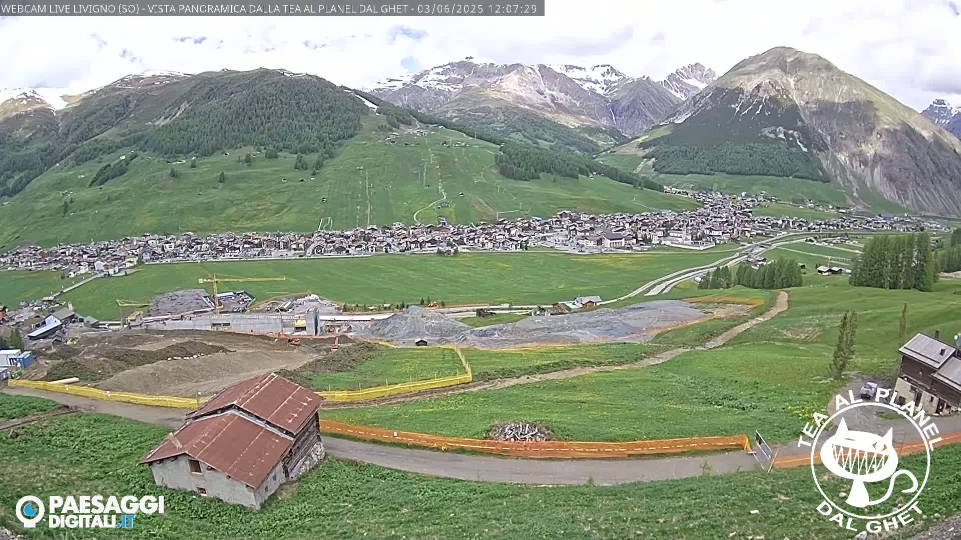 A mountain village nestled in a valley, with ongoing construction visible in the foreground, under a partly cloudy sky.