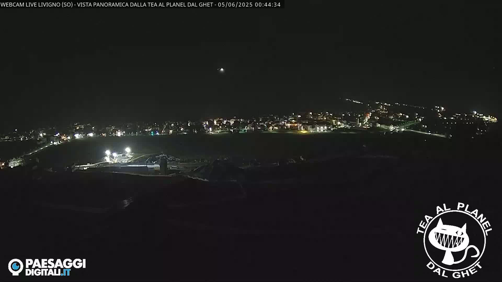 A nighttime, long-range view of a town's illuminated buildings and structures, situated on a slightly elevated plain.