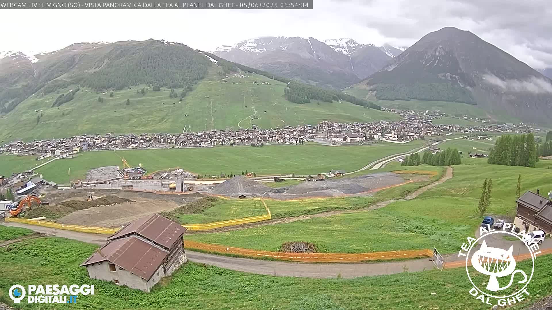 A mountain village nestled in a valley with overcast skies, construction visible in the foreground, and snow-capped mountains in the background.