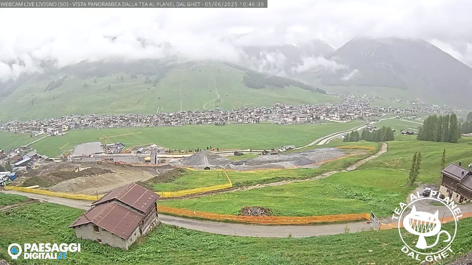 A panoramic view of a town nestled in a valley surrounded by green hills and mountains under cloudy, possibly rainy conditions, shows construction activity and a building near the bottom.