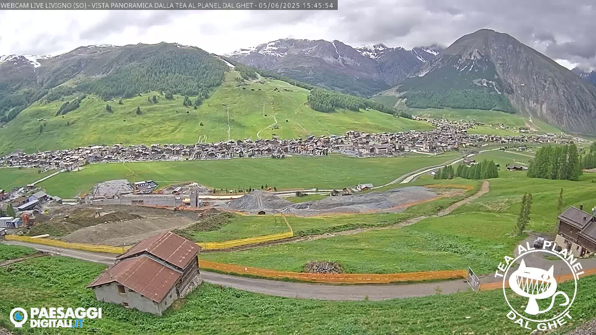 A mountain village nestled in a verdant valley under a partly cloudy sky, with construction visible in the foreground.