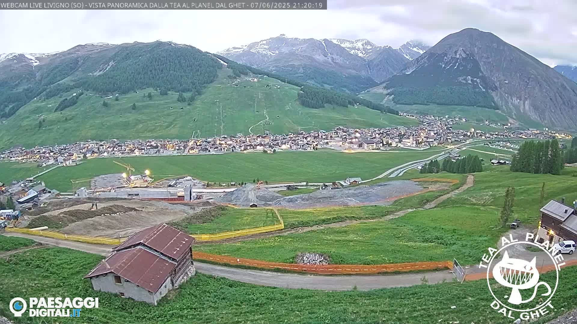 A mountain town at dusk is nestled in a valley below snow-capped mountains under an overcast sky, with construction visible in the foreground.