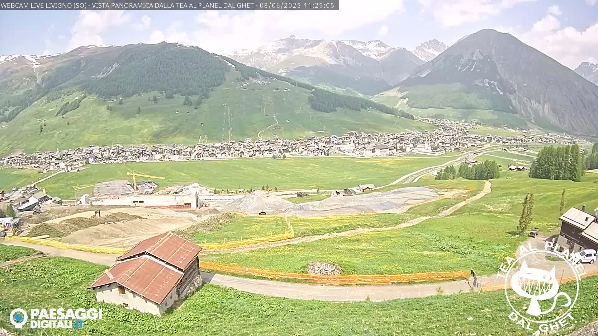 A sunny view of a town nestled in a green valley, surrounded by mountains, with some construction visible in the foreground.