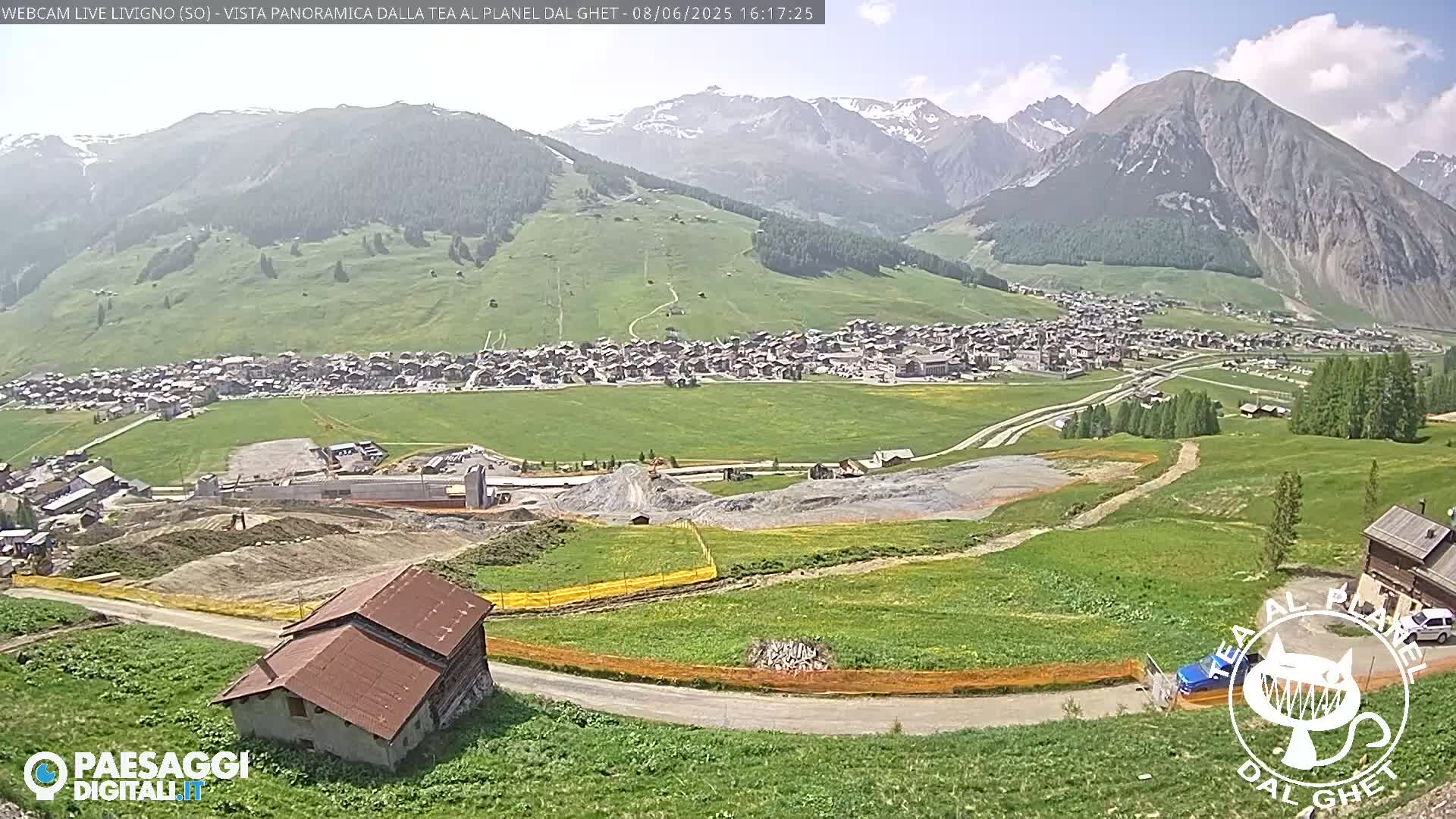 A sunny mountain valley town is visible from a high vantage point, with construction activity and green fields in the foreground.