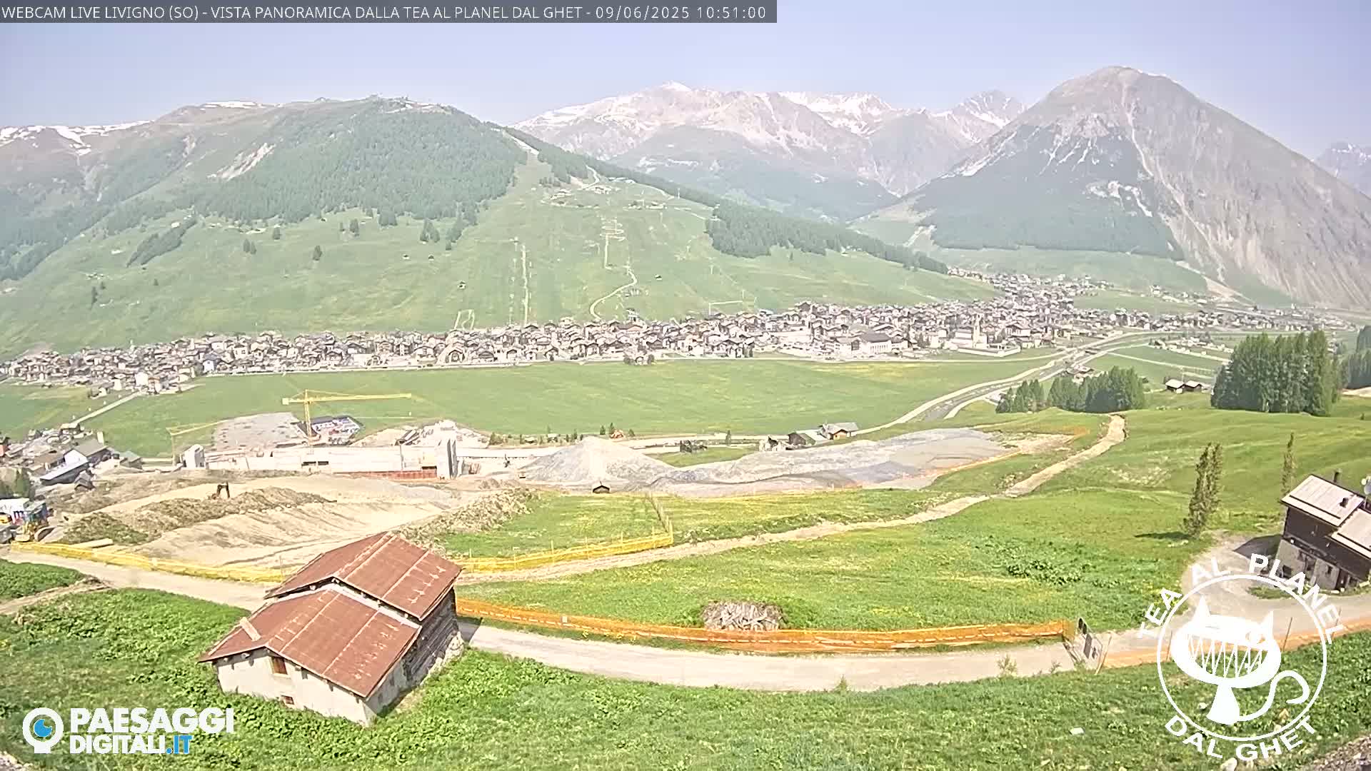 A mountain village nestled in a valley, with construction visible in the foreground, under a clear sunny sky.