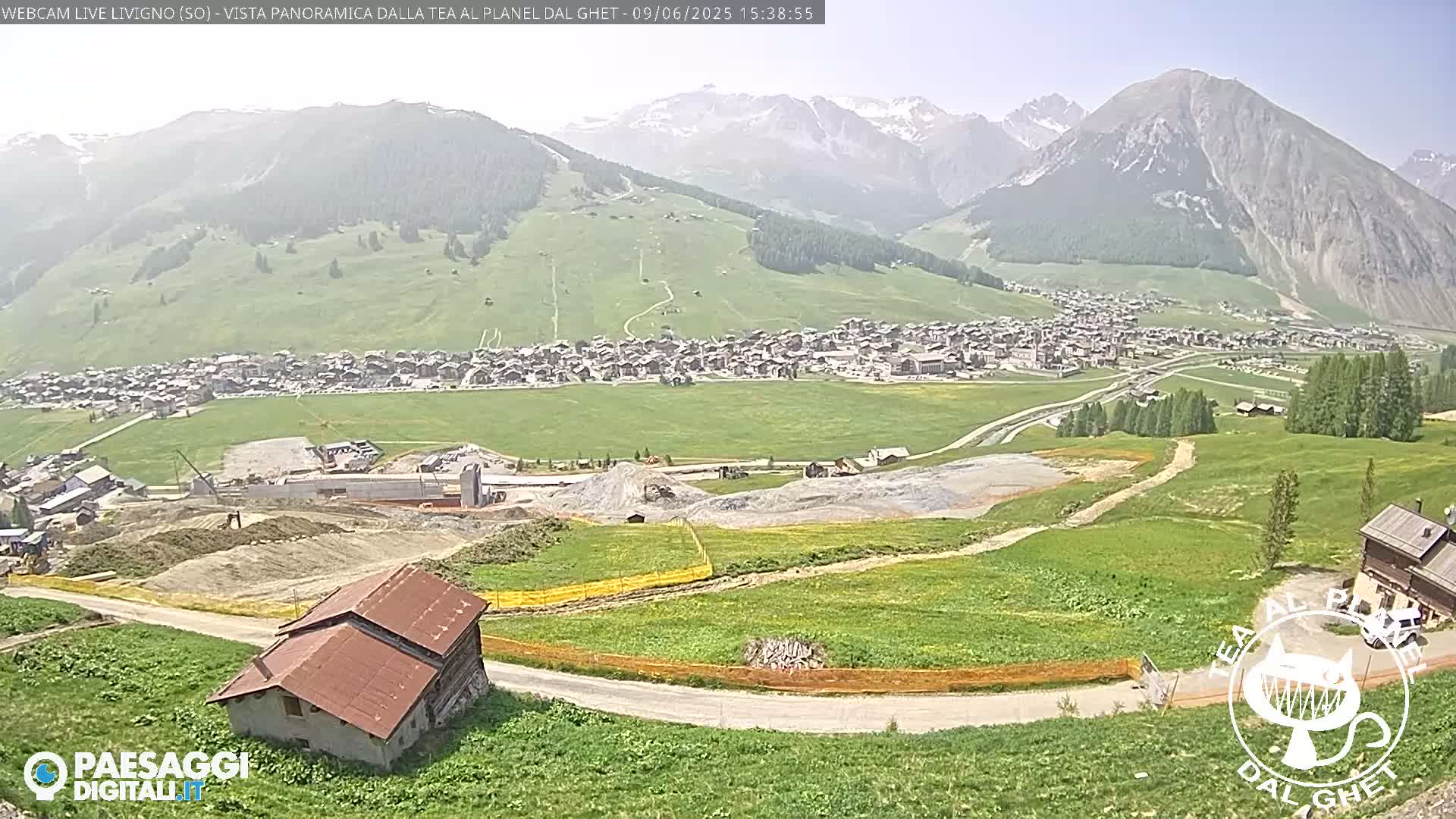 A sunny, panoramic view of a mountain village with green fields, construction sites, and a few buildings, all nestled in a valley beneath snow-capped mountains.