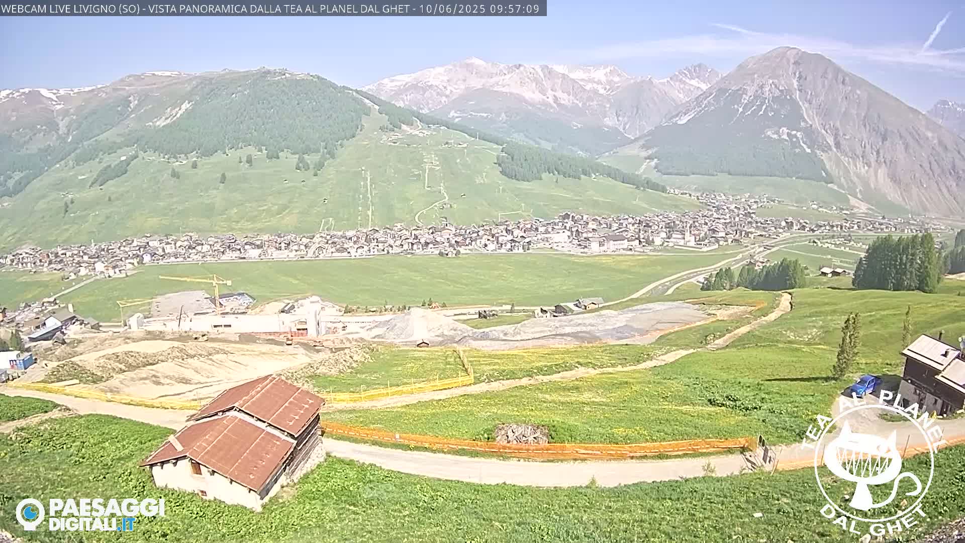 A mountain town nestled in a valley under a sunny sky, with construction visible nearby and a small building in the foreground.