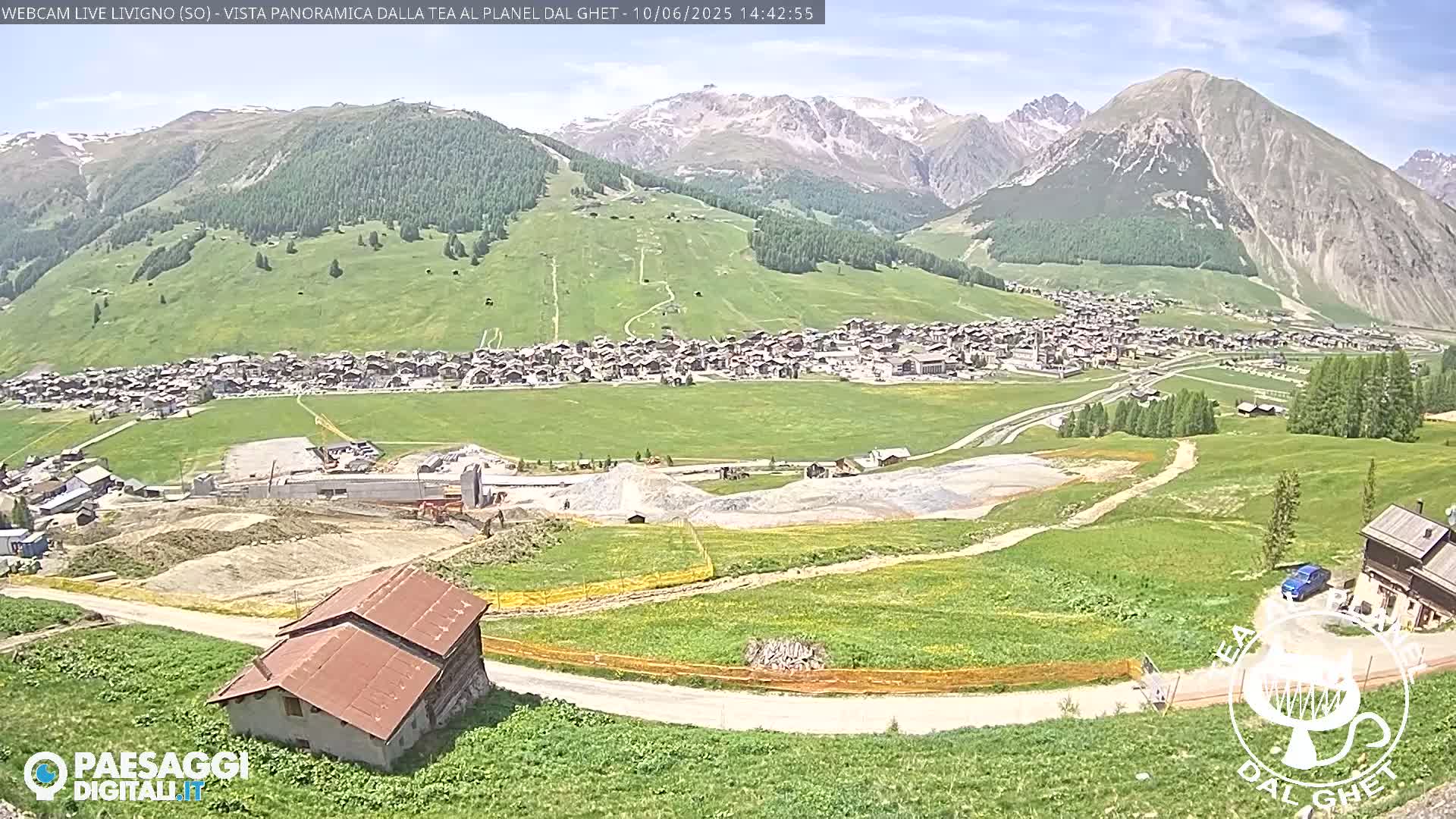 A sunny view of a town nestled in a valley surrounded by green hills and mountains under a mostly clear sky.