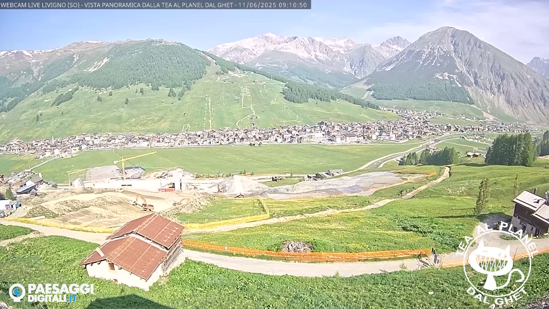 A sunny mountain village is nestled in a valley, with construction visible in the foreground and snow-capped mountains in the background.