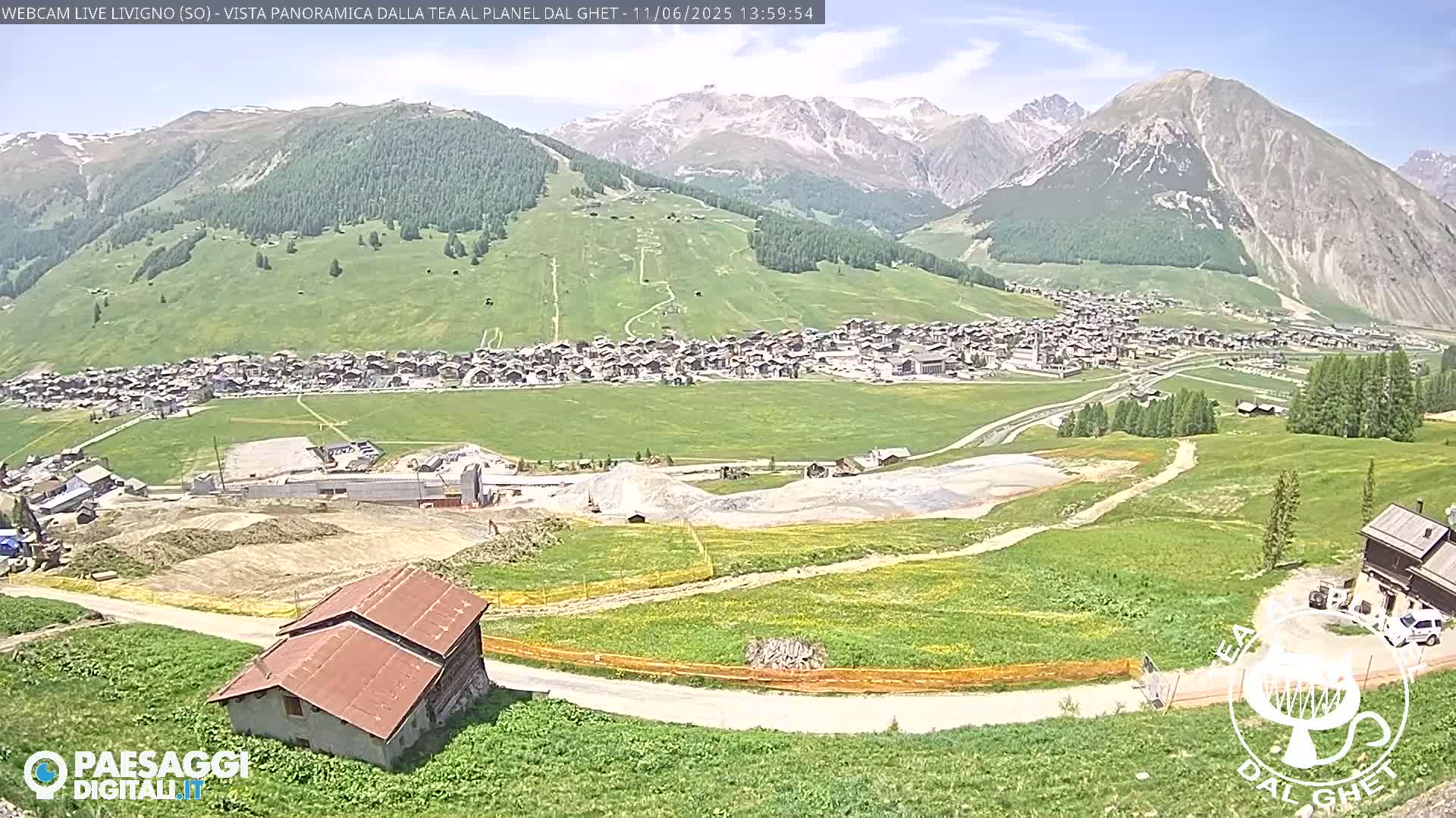 A sunny, mountainous landscape shows a village nestled in a valley, with construction activity visible near a dirt road leading to a small building.