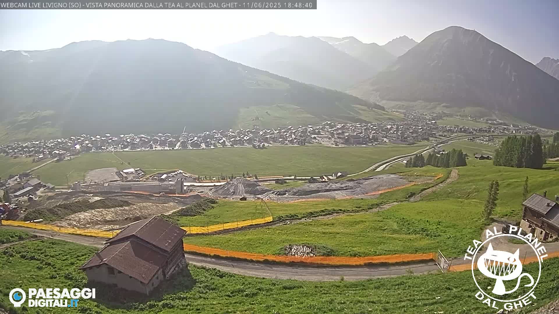 A hazy, sunny day reveals a mountain village nestled in a valley, with ongoing construction visible below, surrounded by green hills and mountains.