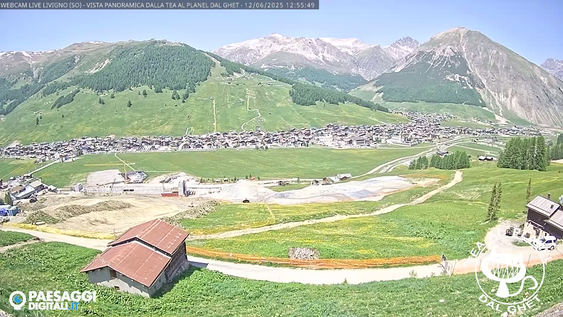 A sunny, high-altitude village nestled in a green valley, surrounded by mountains with some snow-capped peaks, shows construction activity in the foreground.
