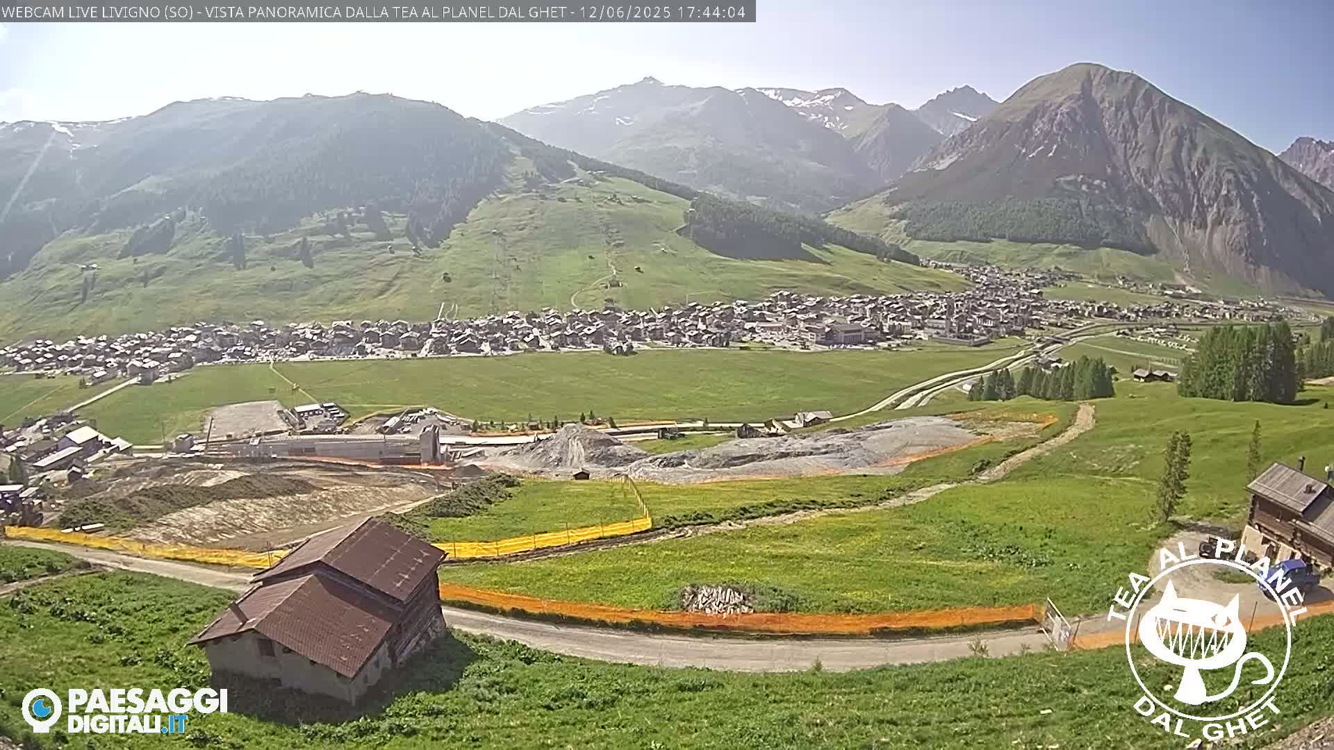 A sunny, high-altitude village nestled in a green valley, surrounded by mountains, with some ongoing construction visible in the foreground.