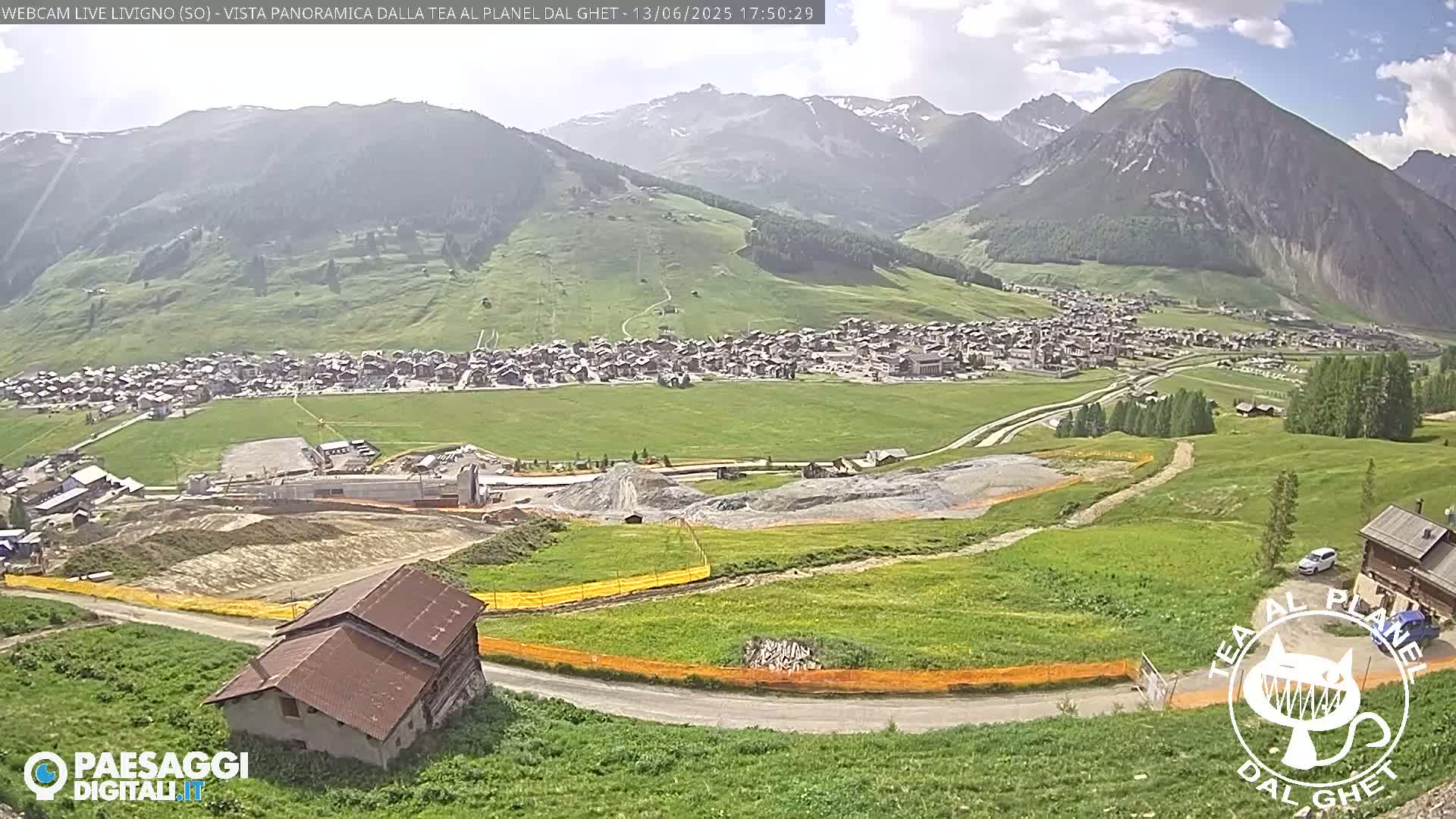 A partly sunny view of a mountain village nestled in a valley, with construction visible in the foreground and green hillsides stretching towards hazy mountains in the background.