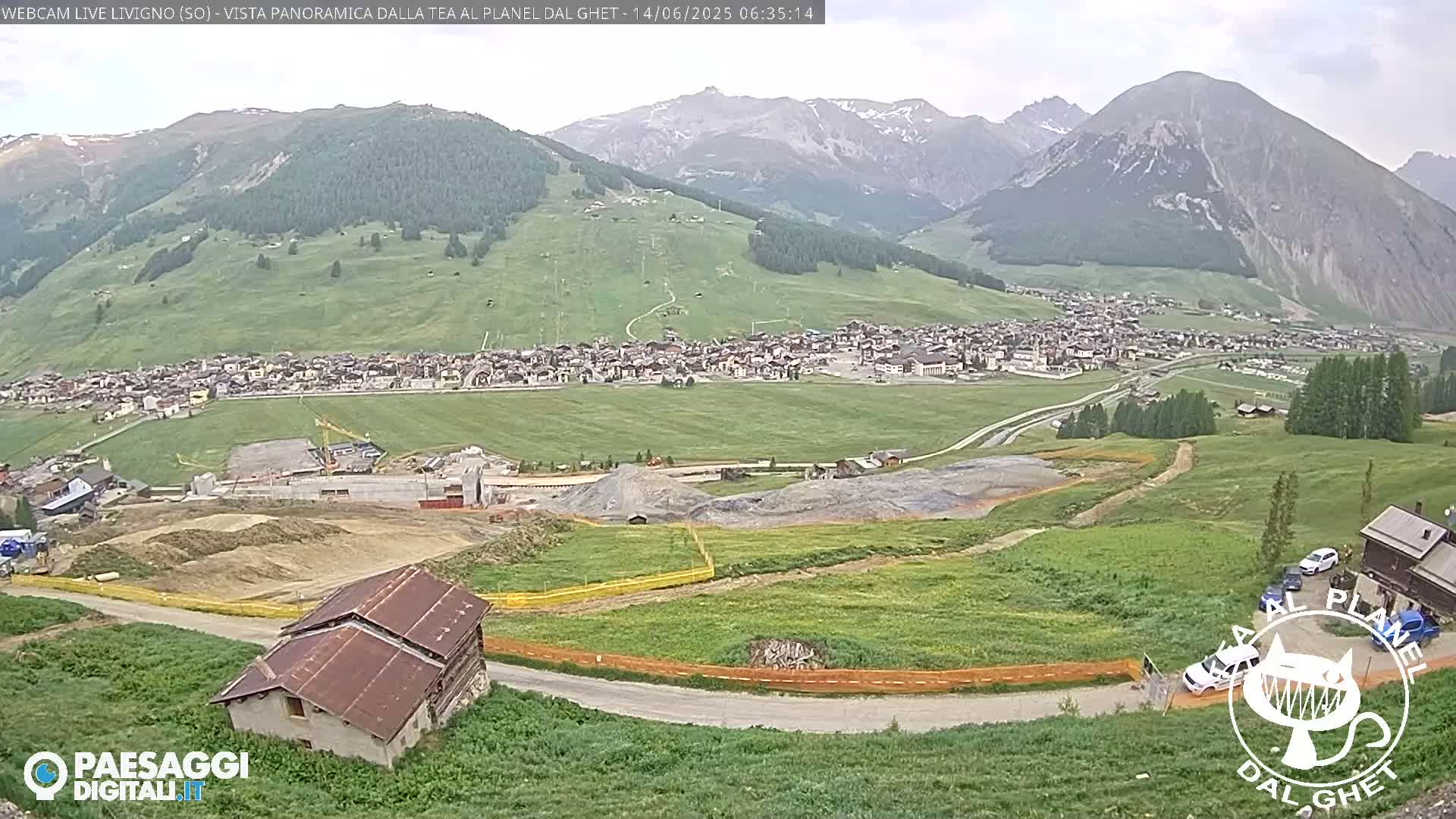 A partly sunny view of a mountain village nestled in a valley, with construction visible in the foreground and green hillsides stretching towards hazy mountains in the background.