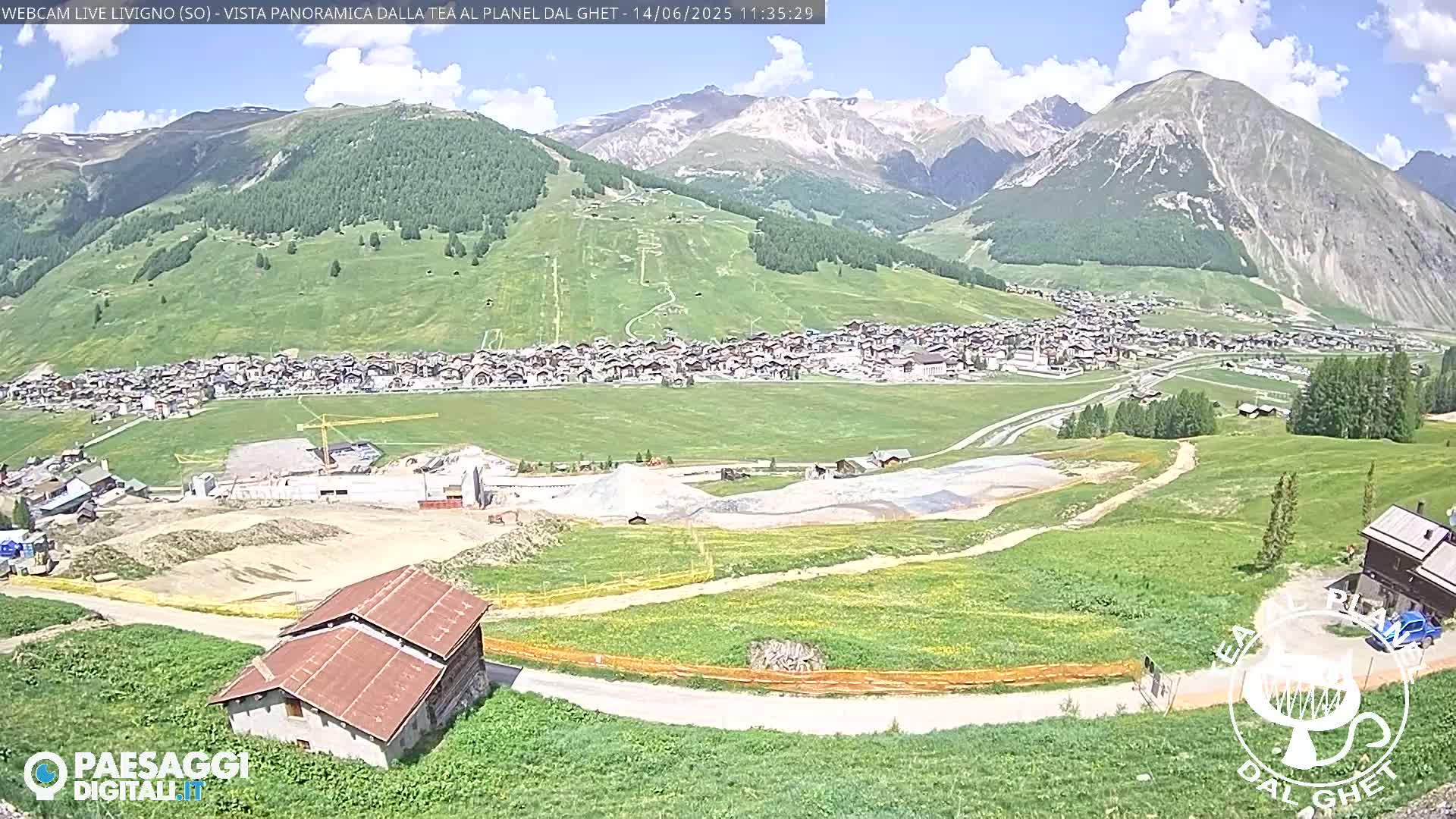 A partly sunny day reveals a mountain village nestled in a valley, with construction visible in the foreground and snow-capped mountains in the background.