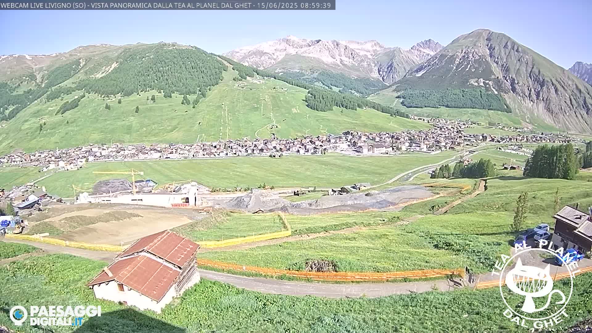 A sunny mountain village nestled in a valley, with construction visible near a small building and grassy fields stretching towards snow-capped peaks in the background.