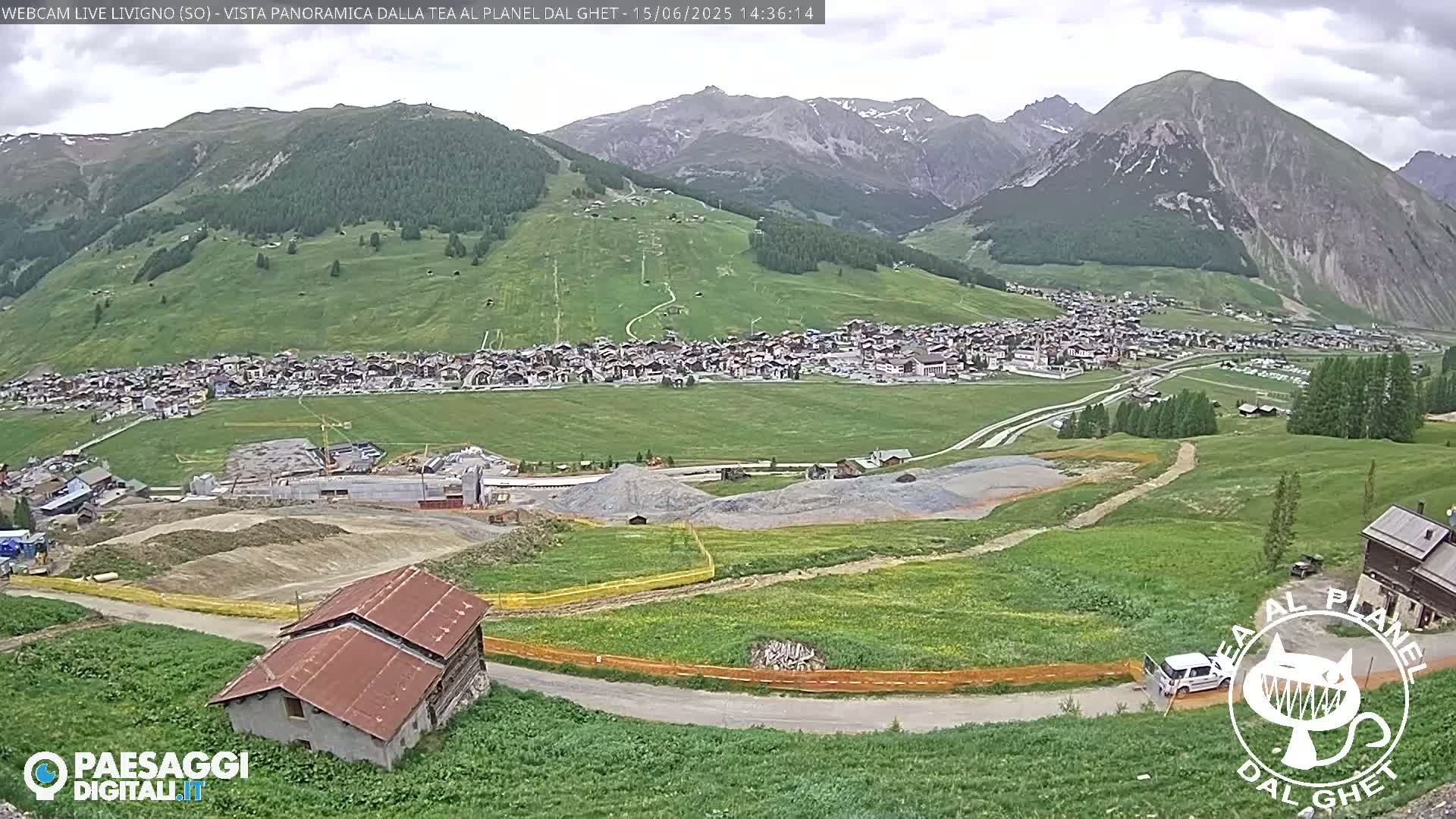 A mountain village nestled in a valley, with green hills and mountains in the background under an overcast sky, shows construction activity near a building.