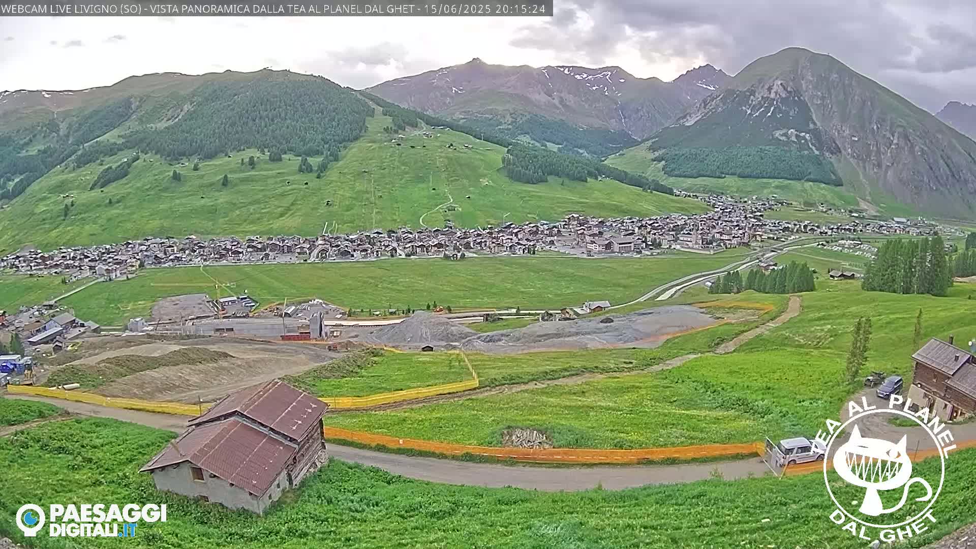 A mountain village nestled in a verdant valley under a partly cloudy sky, with construction visible in the foreground.