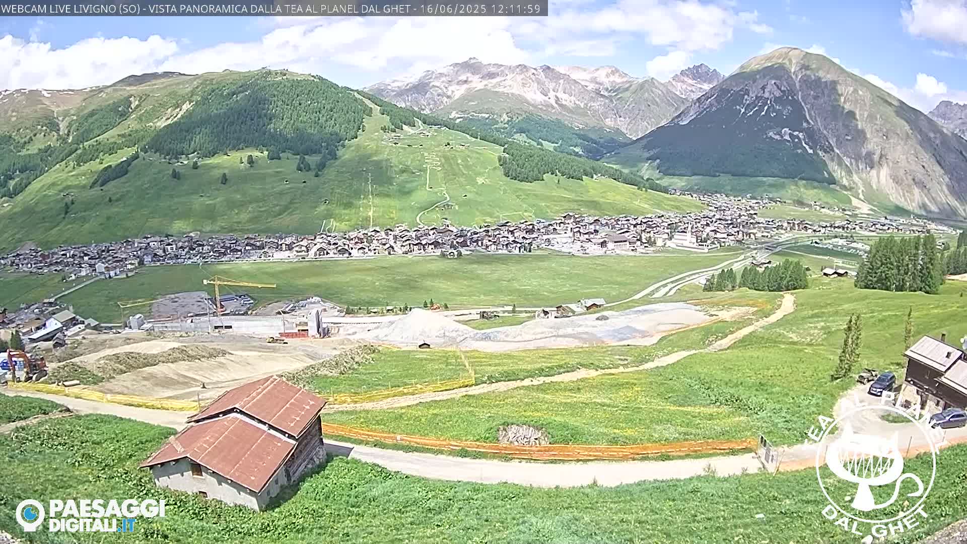 A sunny mountain village nestled in a valley, with construction visible in the foreground and snow-capped mountains in the background.